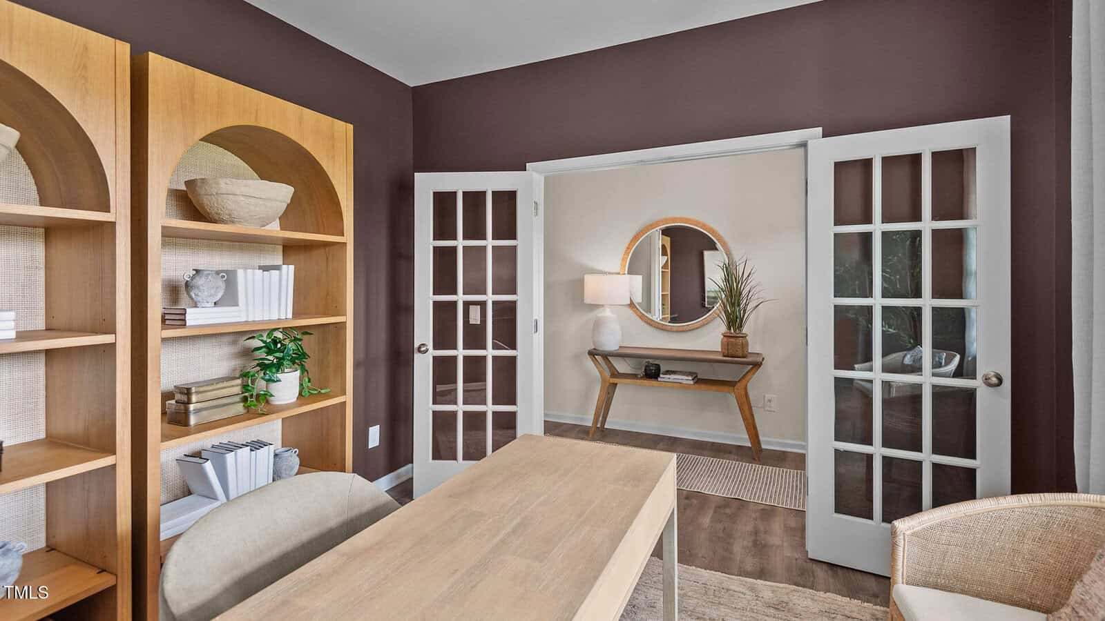 A home office with a wooden desk, beige chair, built-in wooden shelves with books and decor, glass French doors, and a hallway with a round mirror and console table visible in the background.