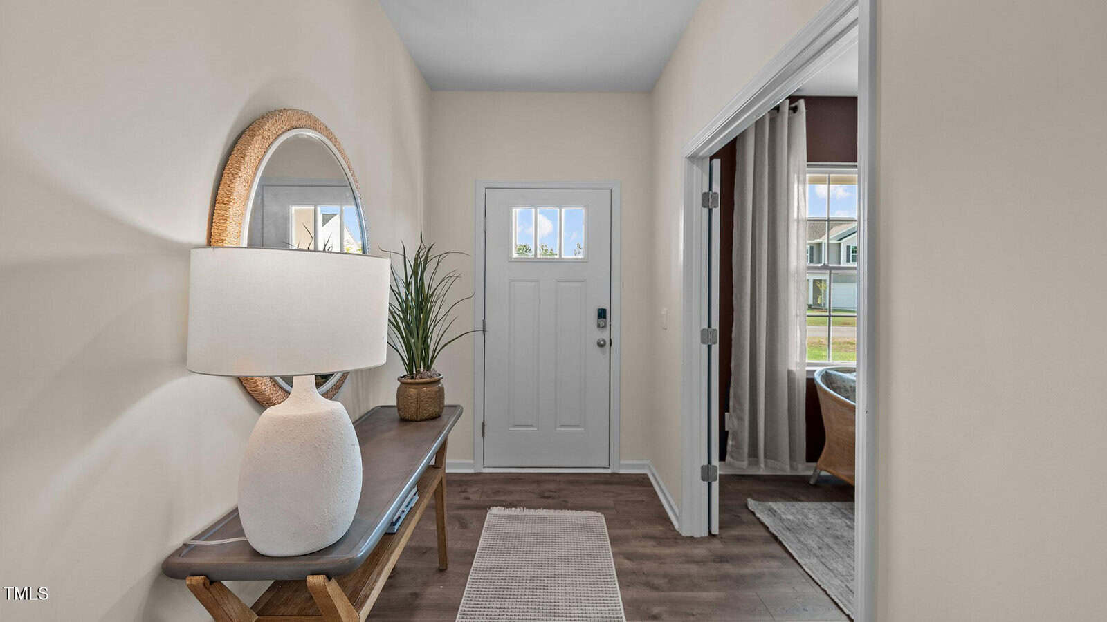 A small entryway with a white front door, a wooden console table with a large round mirror, a white lamp, a potted plant, and a textured rug on a wood floor. A window shows a view of houses outside.
