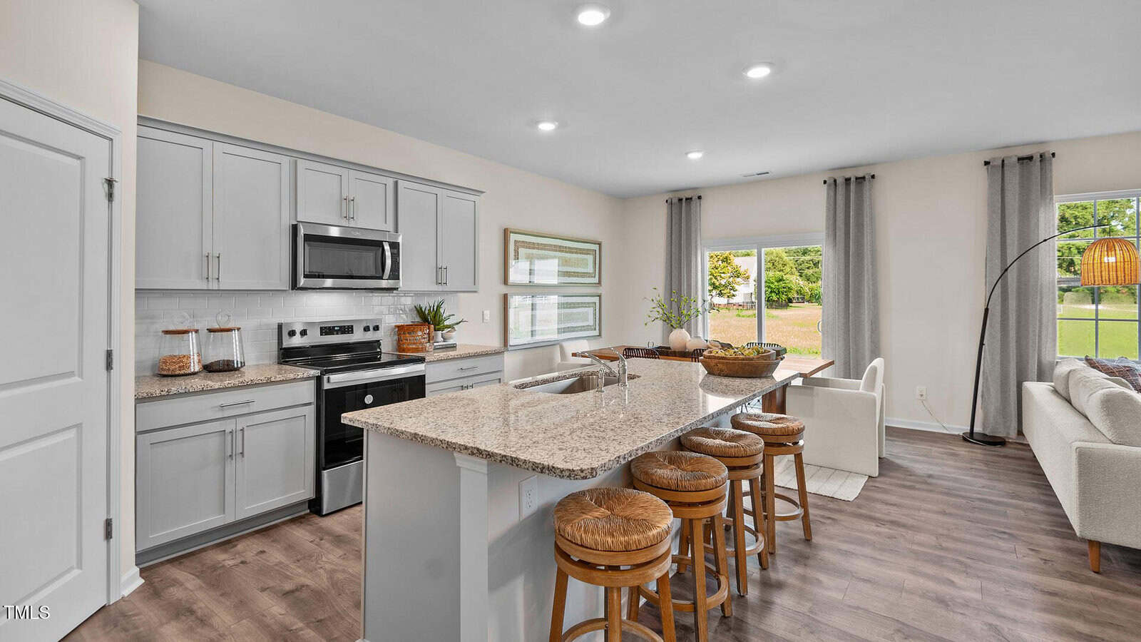 A modern kitchen with light gray cabinets, stainless steel appliances, a granite island with three wooden barstools, and an open layout connecting to a living area with large windows and a white sofa.