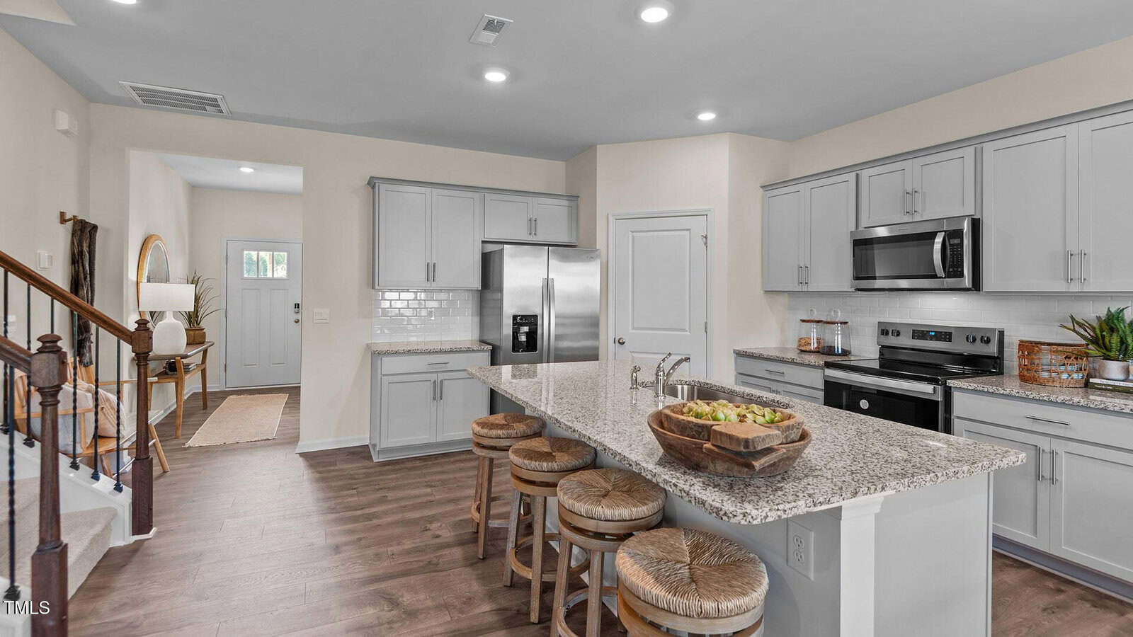 Modern kitchen with light gray cabinets, stainless steel appliances, a granite island with four wooden stools, a fruit bowl centerpiece, and wood flooring. The entryway and staircase are visible in the background.