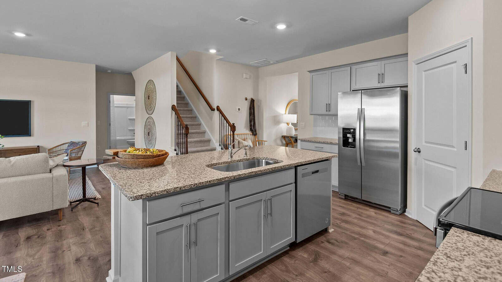Modern kitchen with gray cabinets, granite countertops, stainless steel appliances, and a central island with a sink. Open layout leads to a living area, staircase, and a hallway. Wood flooring throughout the space.