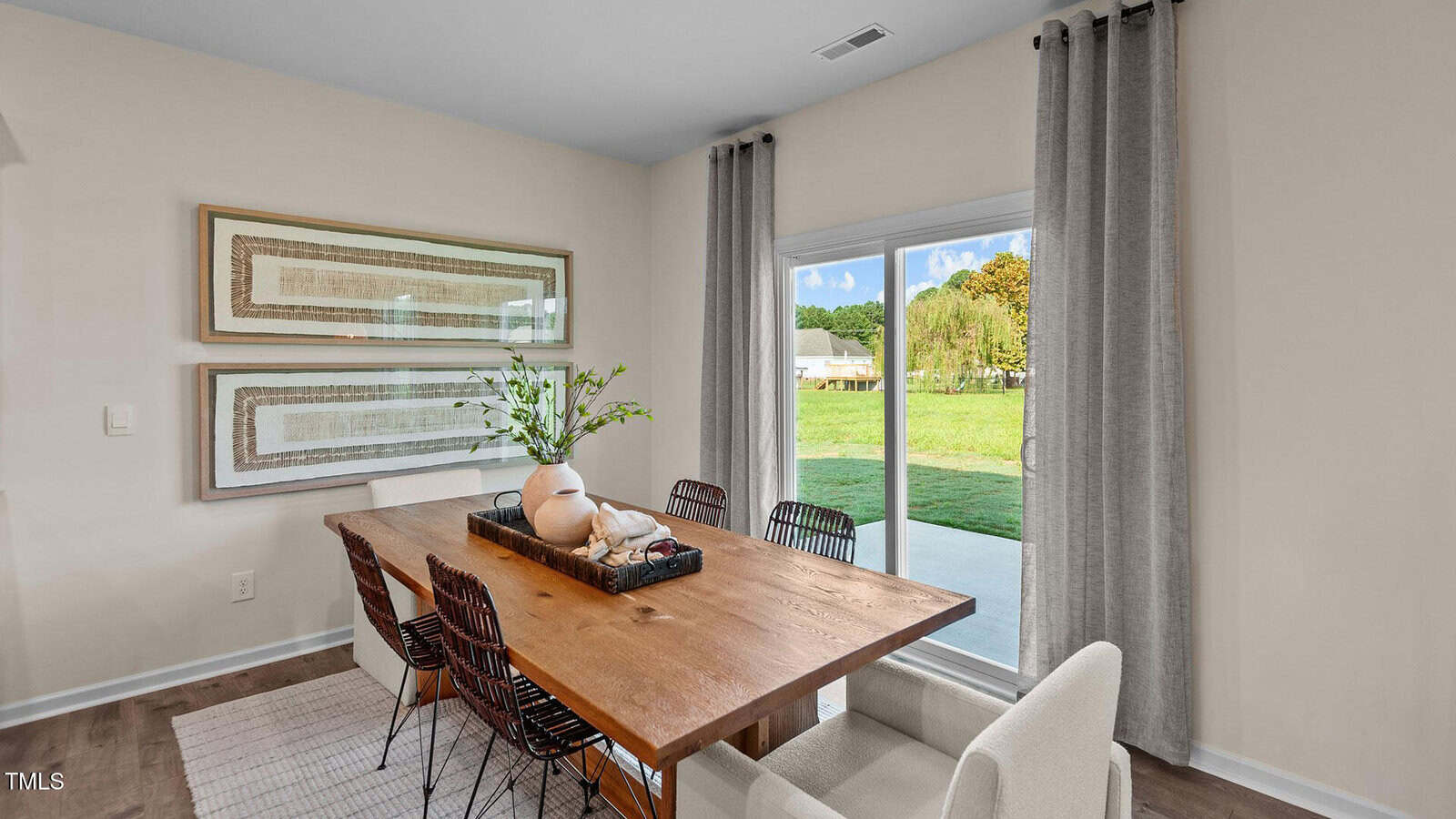 A dining room with a wooden table, six chairs, a vase with greenery, and two framed artworks on the wall. Large window with gray curtains overlooks a green yard.