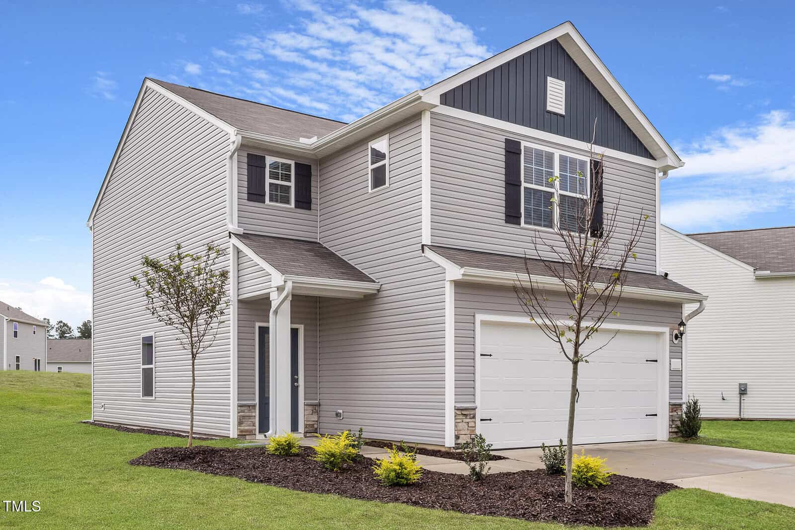 A two-story house with gray siding and a white garage door graces 108 Scoville Street in Oxford. The front yard boasts a small landscaped area with young trees and shrubs, perfectly complementing the partly cloudy sky.