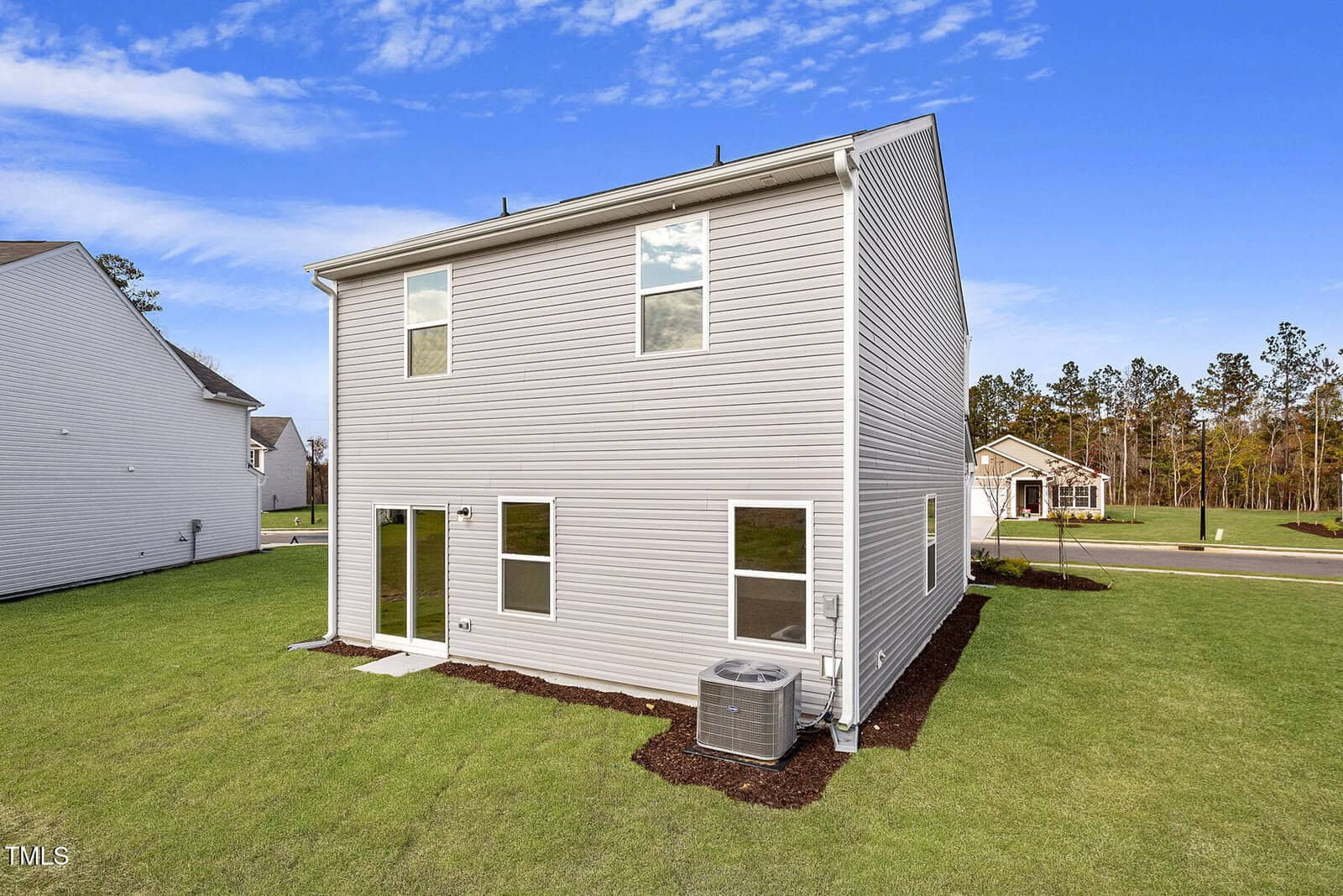 A two-story house with light gray siding and a sloped roof. The backyard features a small, well-maintained lawn, an air conditioning unit, and sliding glass doors. The sky is clear with a few clouds, and a neighboring house is visible.