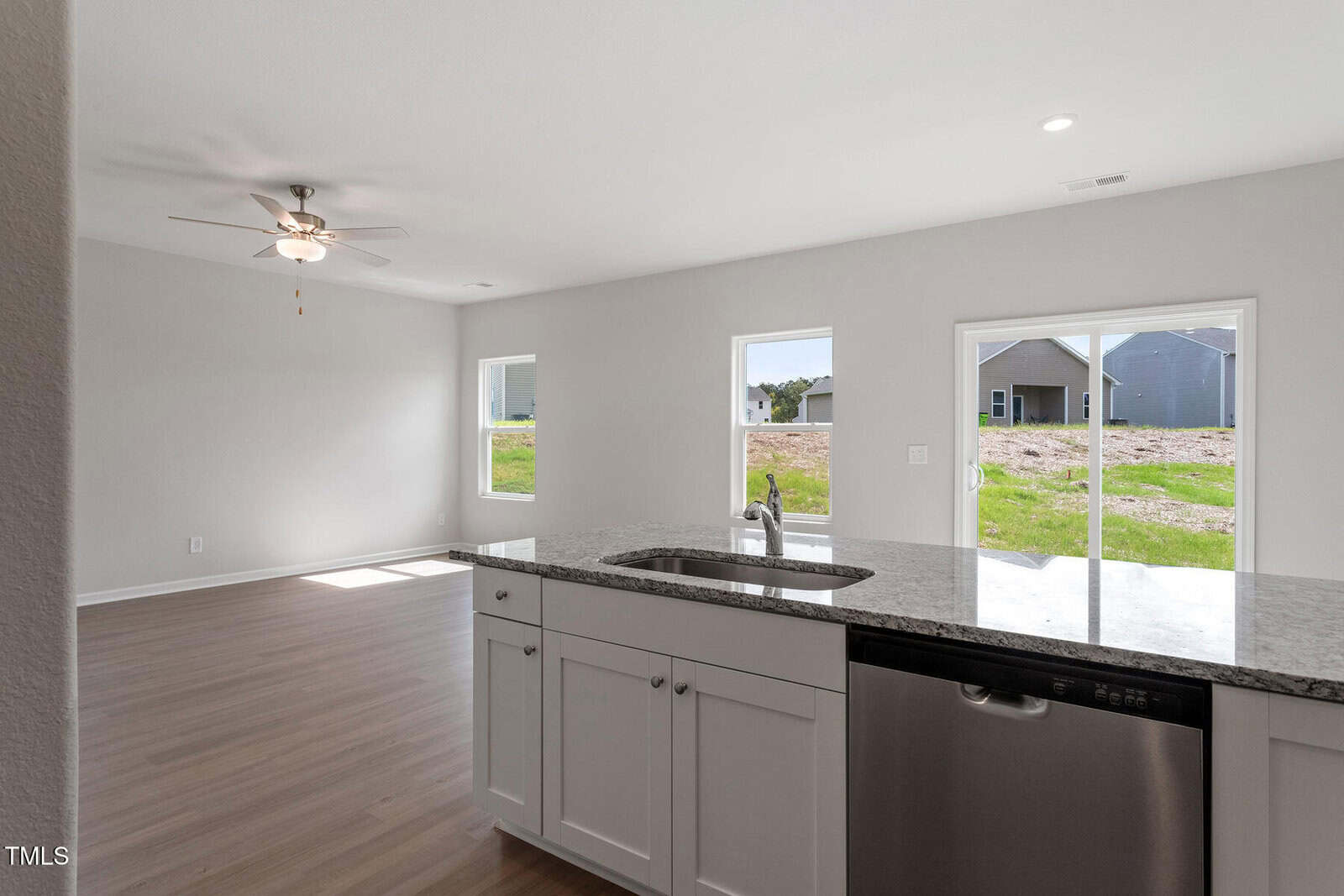 A modern, unfurnished interior of a house with an open floor plan. The image shows a kitchen area with white cabinets, a granite countertop, and stainless steel appliances, including a dishwasher. Large windows offer views of a grassy backyard.