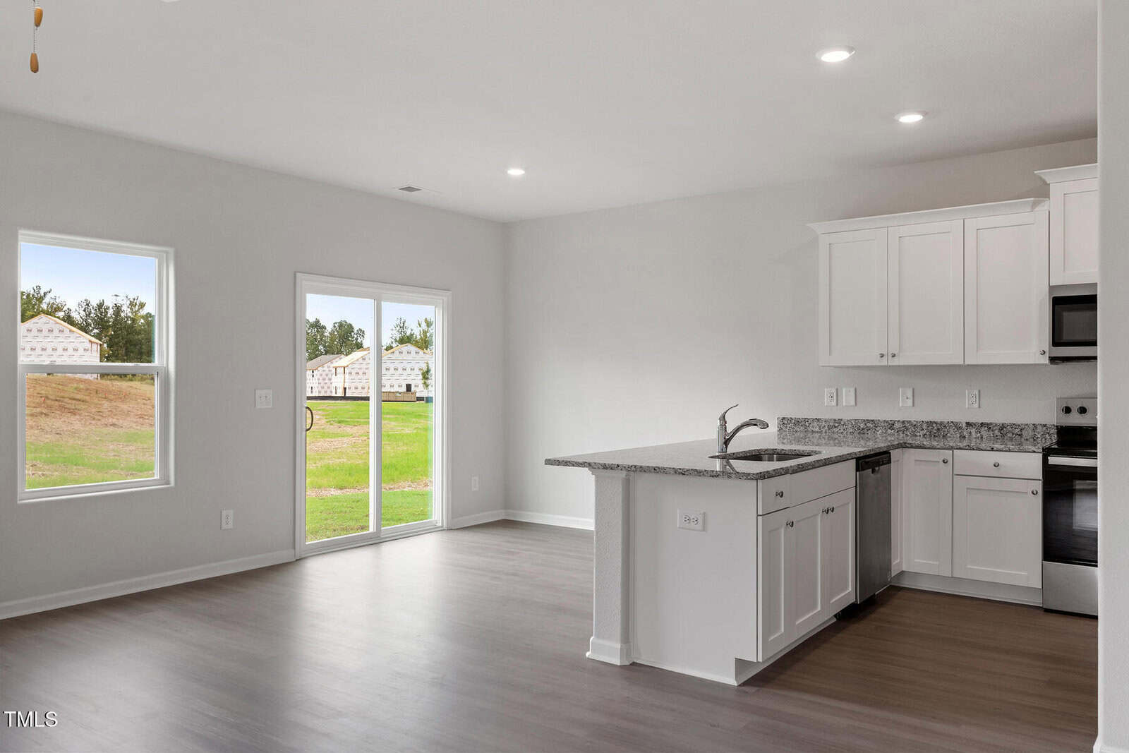 A bright, unfurnished kitchen and living area with light wood flooring. The kitchen features white cabinets, a granite countertop, and stainless steel appliances. A sliding glass door and windows offer a view of the grassy backyard.