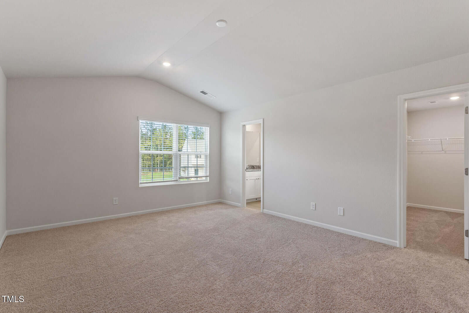 A spacious, empty carpeted room with a large window allowing natural light. The room has white walls and a high ceiling. A door on the right leads to a closet and an adjoining room with a bathroom visible.
