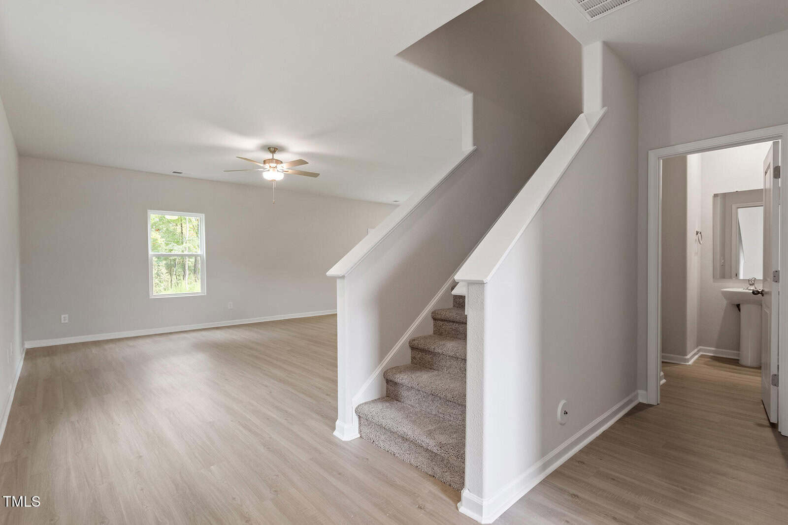 A spacious, empty room with light wood floors and white walls. A staircase with gray carpet leads to the upper level. A ceiling fan is mounted on the ceiling, and there is a small, open door to a bathroom on the right. Natural light enters through a window.