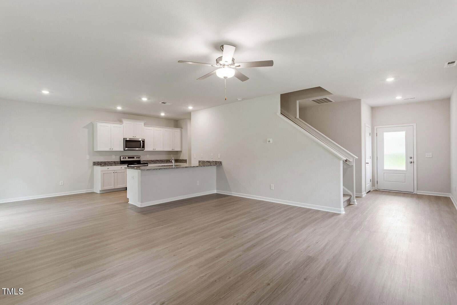 Spacious interior of a modern home with light wood flooring. The open-plan layout includes a kitchen with white cabinets, a center island, and stainless steel appliances. A staircase leads upstairs, and a ceiling fan is mounted on the ceiling.