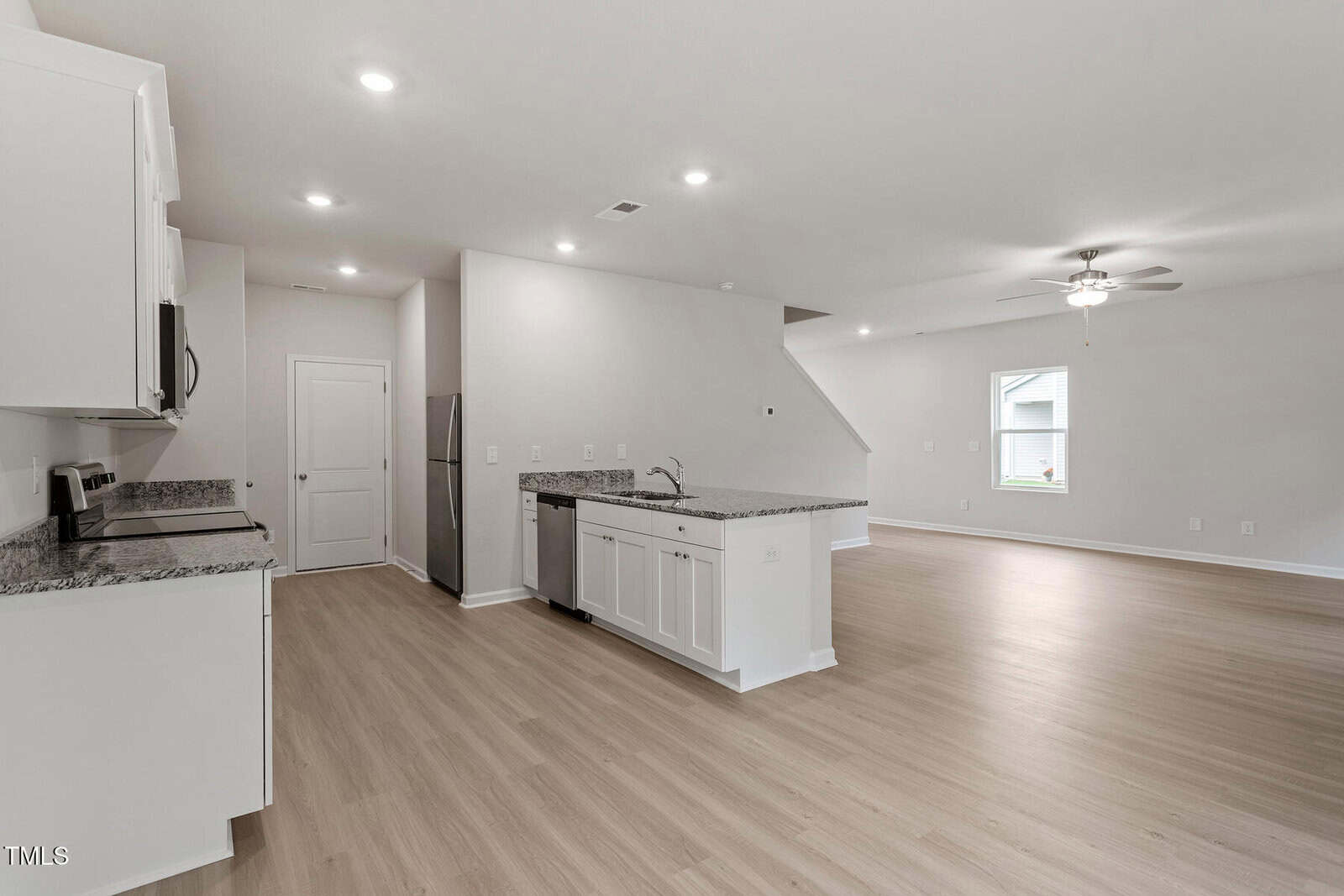 A modern open-concept interior featuring a kitchen with white cabinets, granite countertops, and stainless steel appliances. Adjacent is a spacious living area with light wood flooring, recessed lighting, and a window. A ceiling fan is visible above.