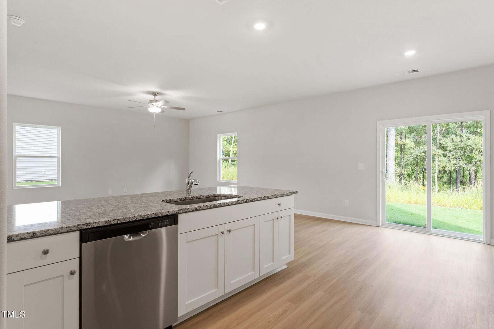 A modern kitchen with white cabinets, a granite countertop island with a built-in sink and dishwasher, opens to a bright living area with light wood flooring. A ceiling fan is installed, and glass sliding doors lead to a grassy outdoor space.