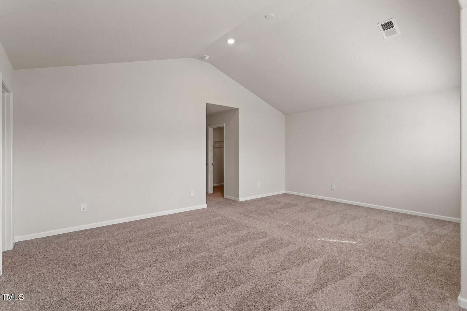 An empty room with beige carpet and light-colored walls. The ceiling is vaulted, and a walk-in closet is visible through an open door. There is a single AC vent on the ceiling and sunlight streaming through a window outside the frame.