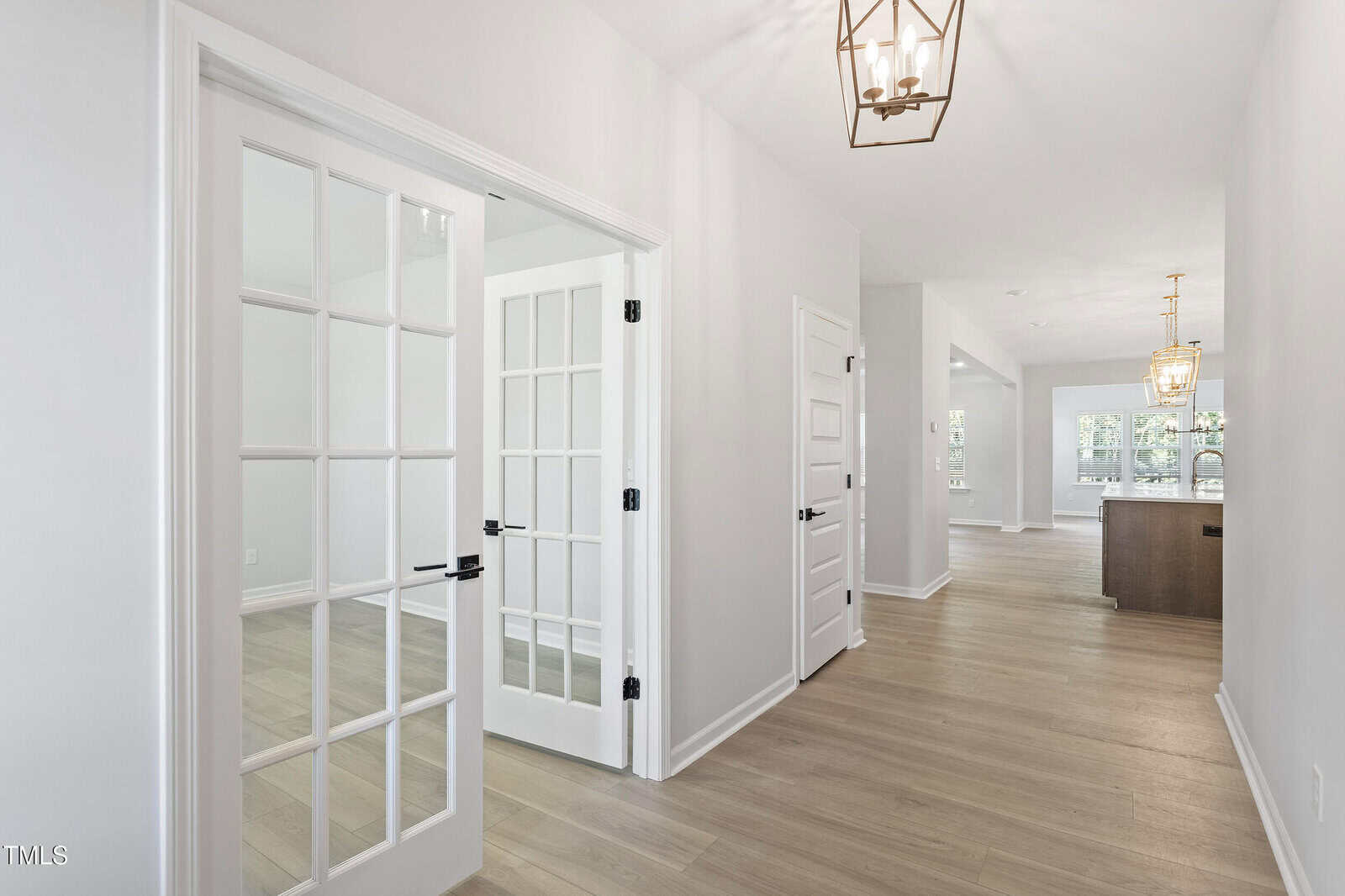 A hallway with light wood floors and white walls features a set of double glass French doors on the left, two white doors further down the hallway, and a kitchen area visible at the far end.