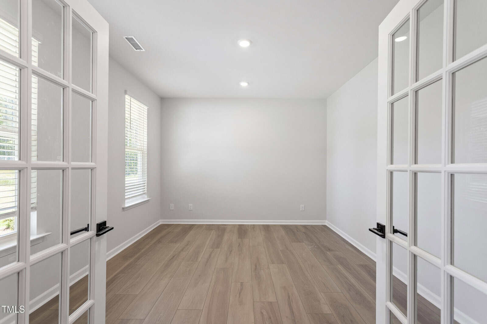 A bright, empty room with light-colored wood flooring, white walls, two large windows on the left, and glass double doors partially open in the foreground. Ceiling lights are turned on.