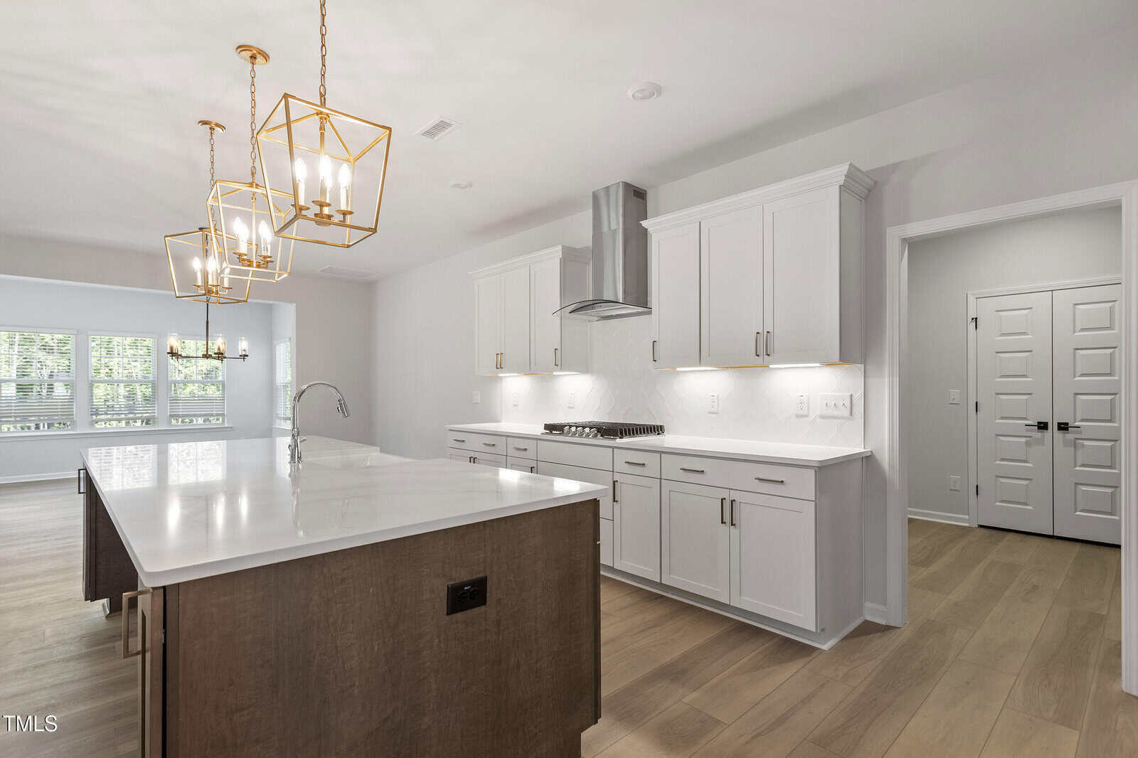 Modern kitchen with white cabinets, stainless steel appliances, large island with marble countertop, pendant lights, light wood floors, and an adjacent dining area with windows. Double doors are visible in the background.