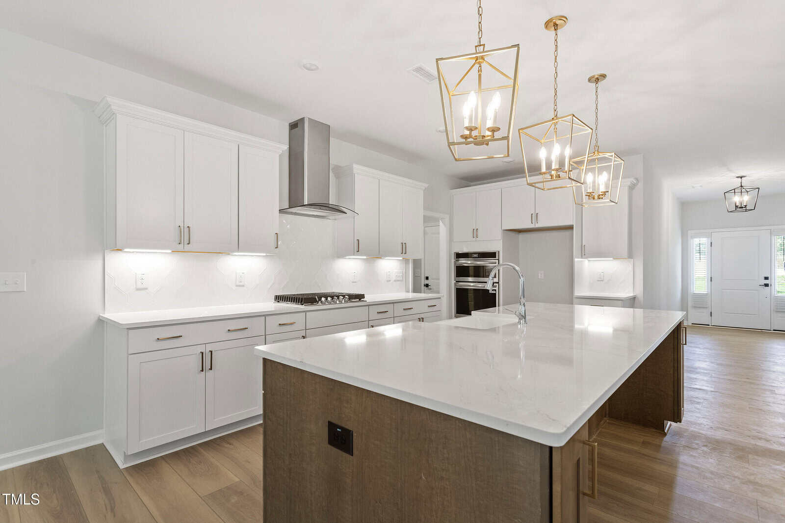 Modern kitchen with white cabinets, stainless steel appliances, a large island with a marble countertop, three gold pendant lights, light wood flooring, and a view toward the entryway with double doors in the background.
