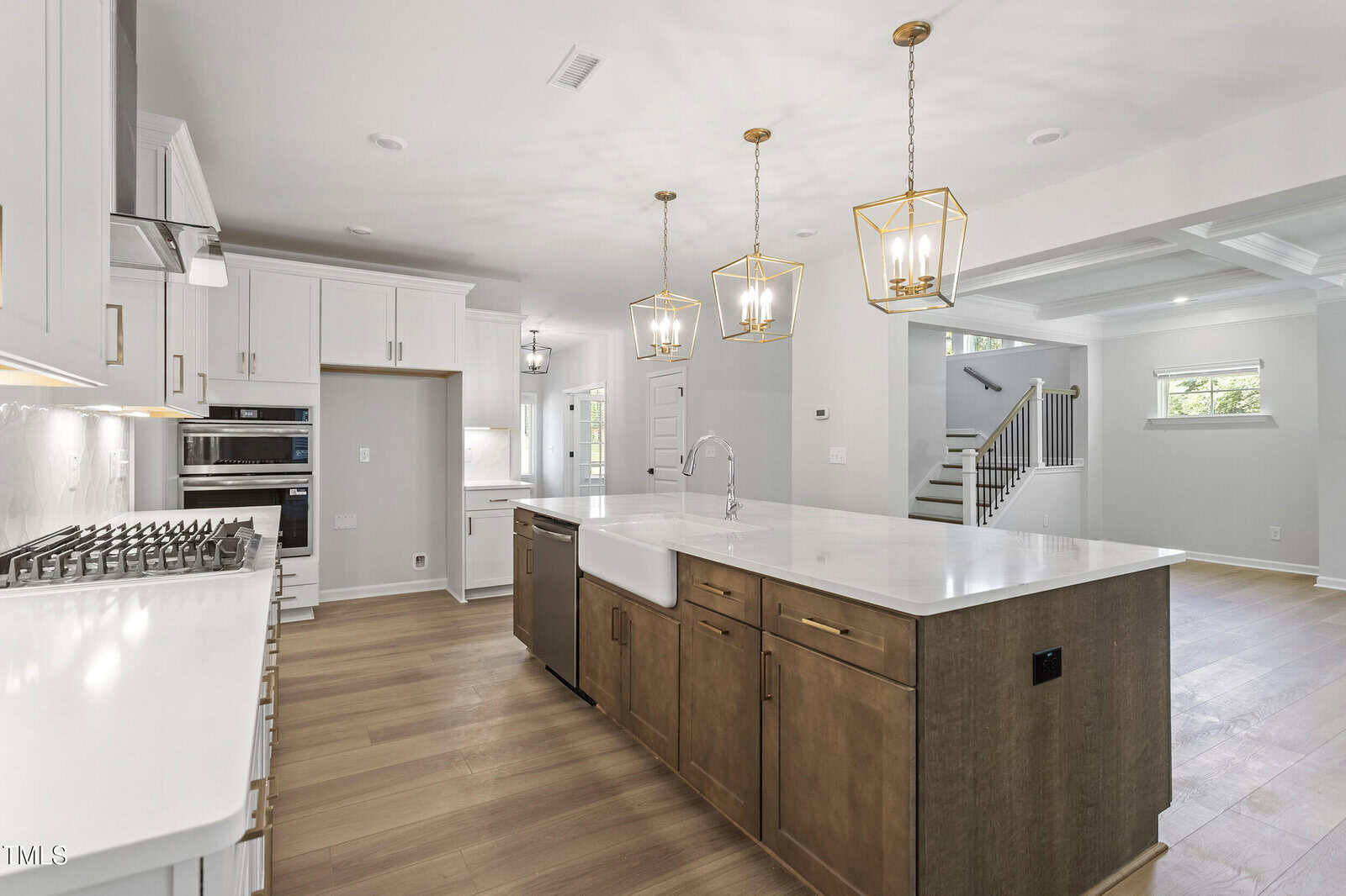 Modern kitchen with white and wood cabinetry, large island with farmhouse sink, three pendant lights above, stainless steel appliances, light wood floors, and view into open living area with staircase.