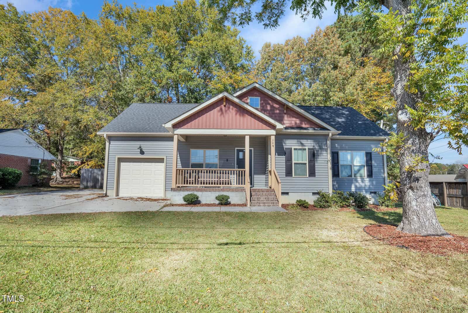 A single-story house with gray siding, a front porch with columns, an attached garage, and a well-maintained lawn. Large trees and other houses are visible in the background.