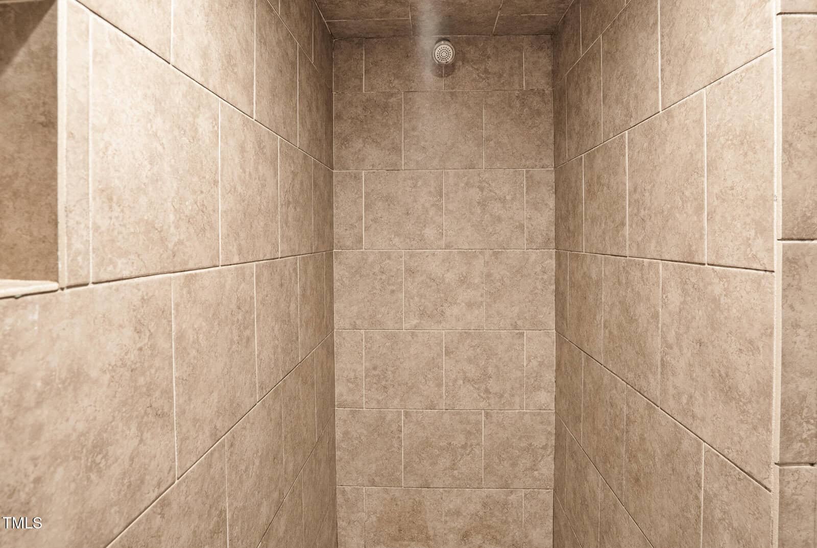 A shower stall with beige ceramic tile walls and ceiling, arranged in a rectangular pattern. A round metal showerhead is mounted on the ceiling. The stall appears empty and well-lit.