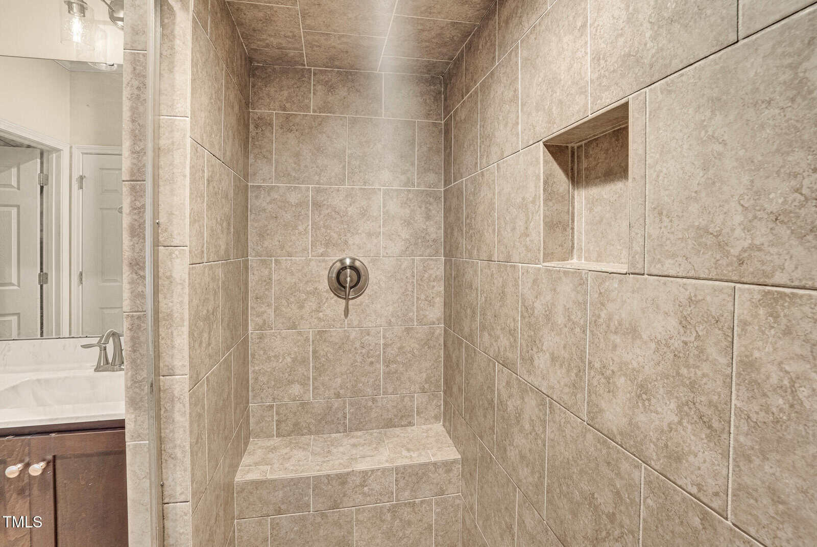 Tiled walk-in shower with a built-in bench, a recessed shelf, and a single central shower control knob. Partial view of a bathroom sink and door is visible outside the shower.