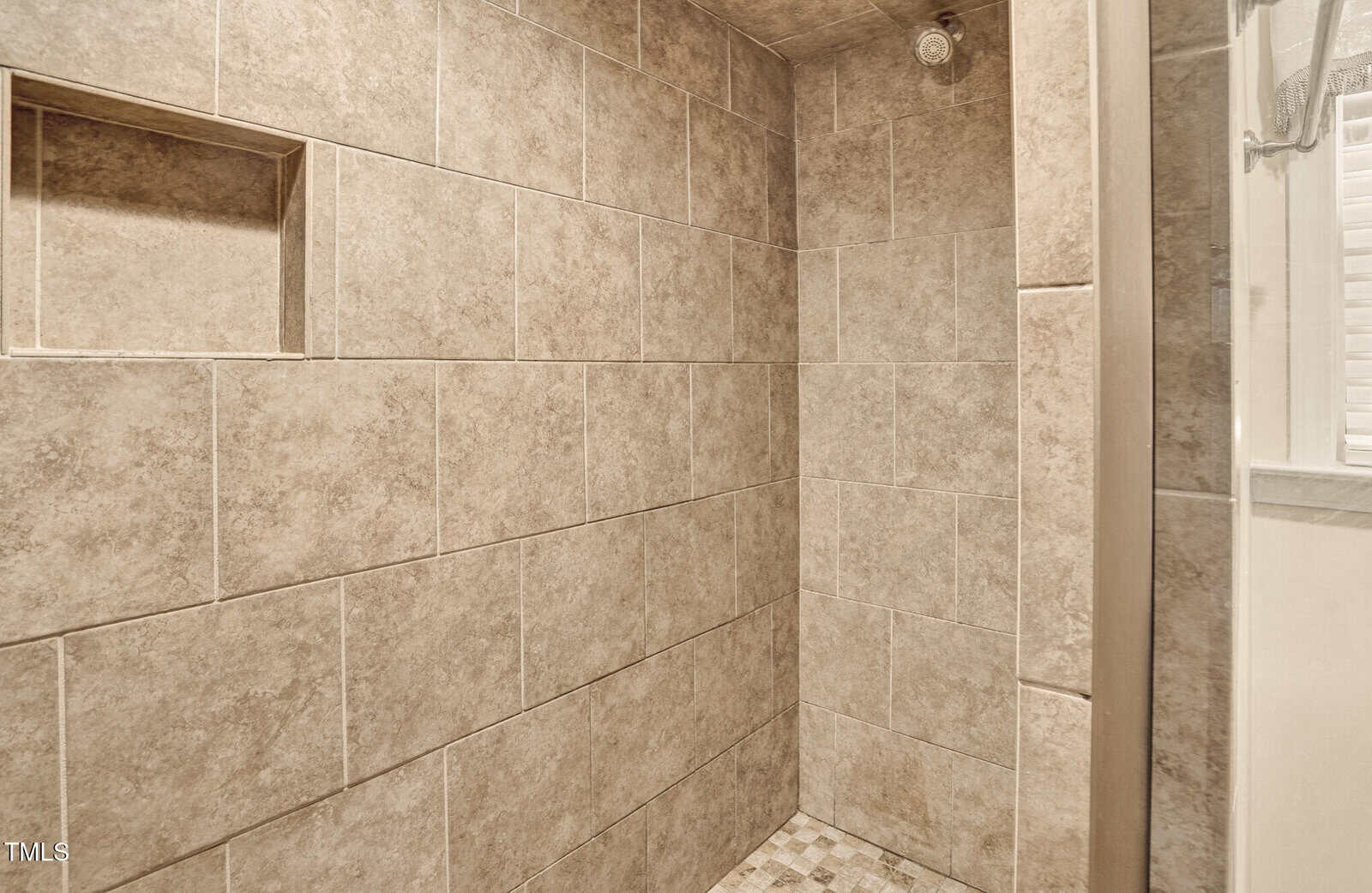 Walk-in shower with beige tile walls and a built-in shelf on the left. The floor is tiled with smaller, square tiles in a checker pattern. A showerhead is mounted on the upper right wall.