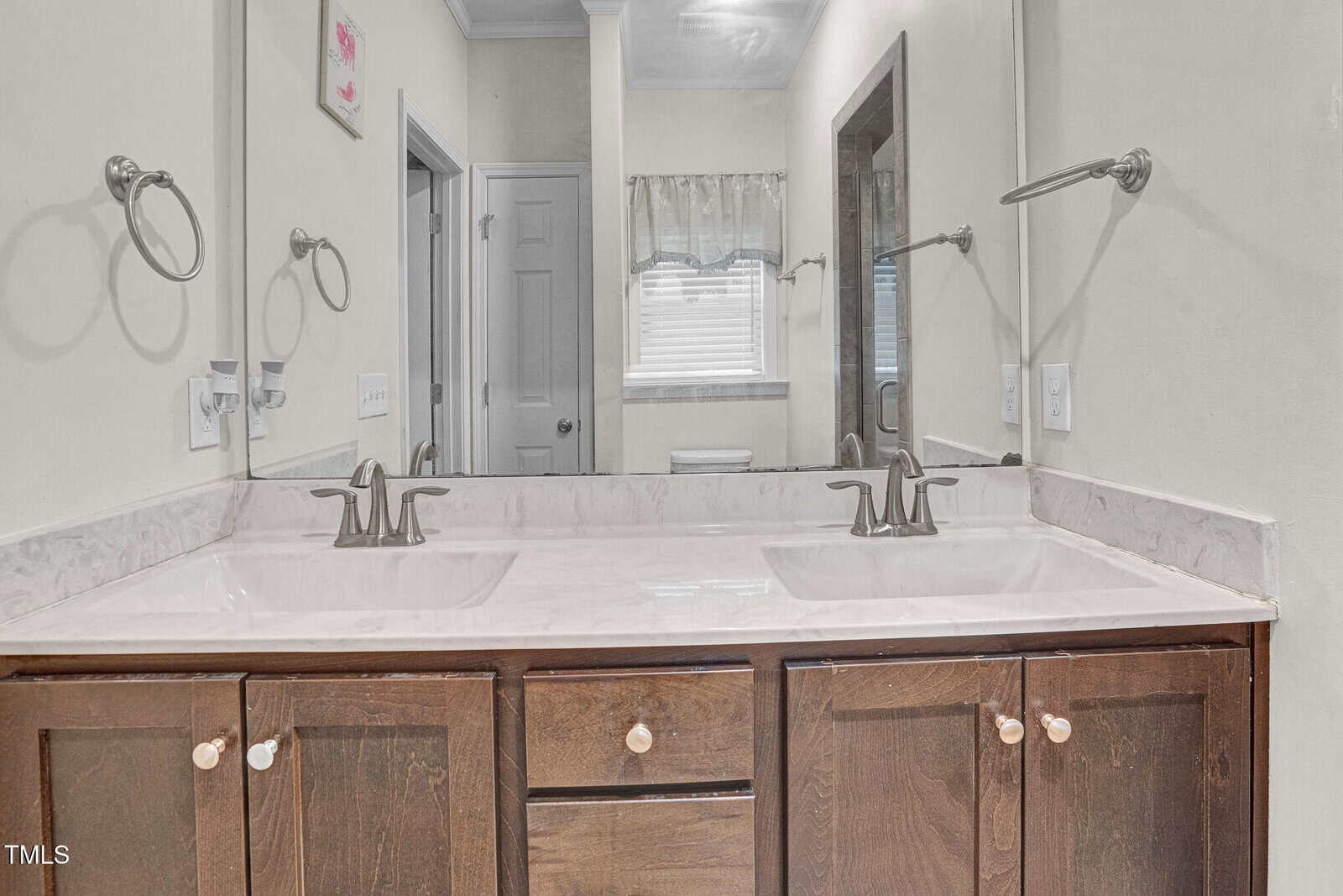 Double sink bathroom vanity with marble countertop, brown cabinets, and two mirrors. Towel rings are mounted on each side. A window and a door are visible in the background, along with a glimpse of a toilet.