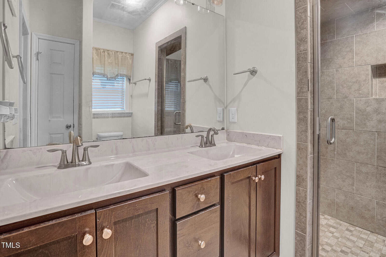A bathroom with a double sink vanity featuring brown wooden cabinets, a large mirror, silver faucets, and a glass-enclosed tiled shower to the right. A window with a beige valance and a door are in the background.