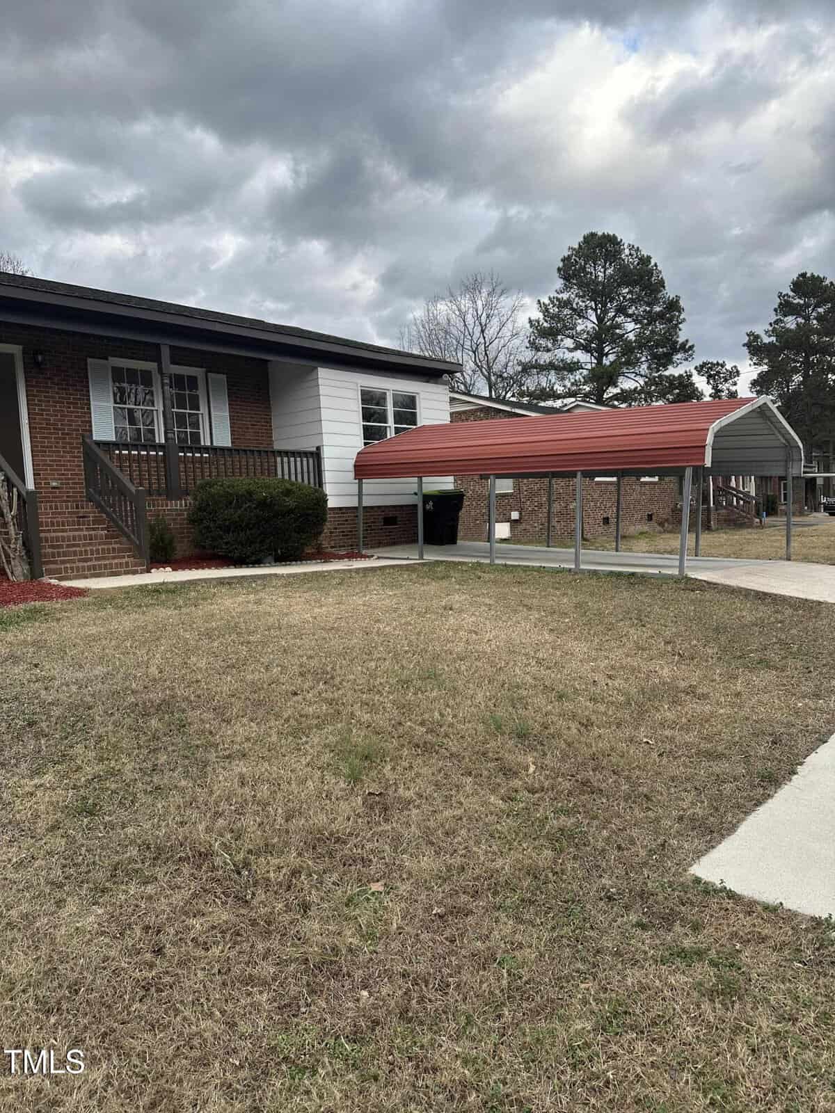 A brick and white siding house with a small front porch is shown. A metal carport with a red roof is situated to the right in the front yard. The lawn is grassy, and there is a concrete path leading to the porch. Cloudy sky in the background.