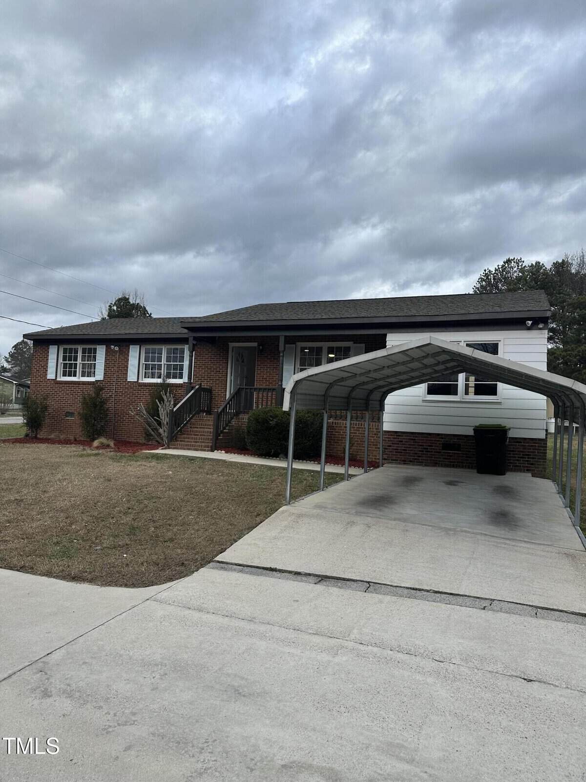 A single-story brick house with a front porch, surrounded by a grassy lawn. The house features a white trim and a covered driveway with a metal carport. The sky is overcast with gray clouds.