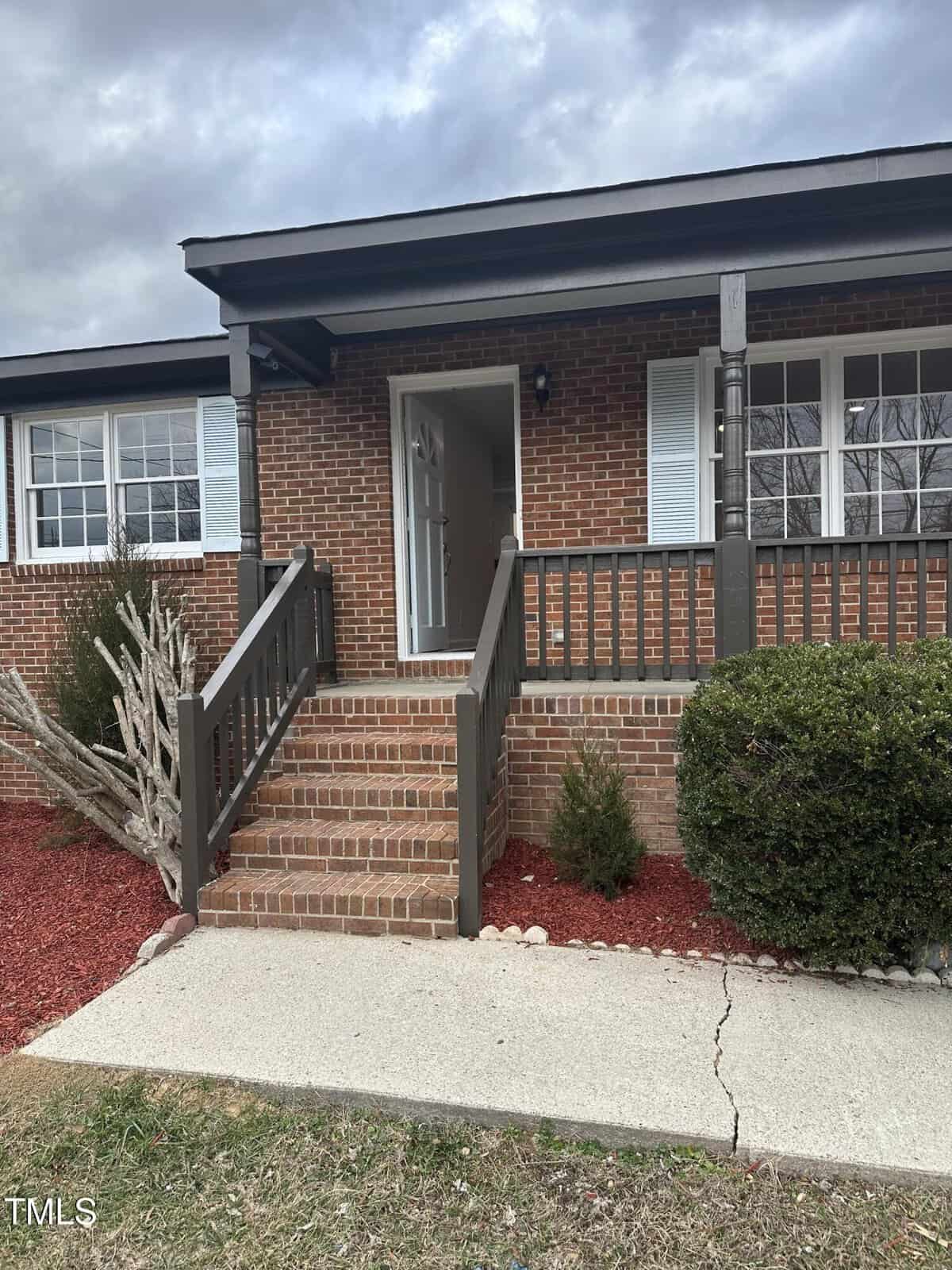 A brick house with a small front porch and wooden railings. The porch is accessed by steps with a pathway leading up to it. Shrubs and red mulch decorate the front yard area. The sky is overcast.