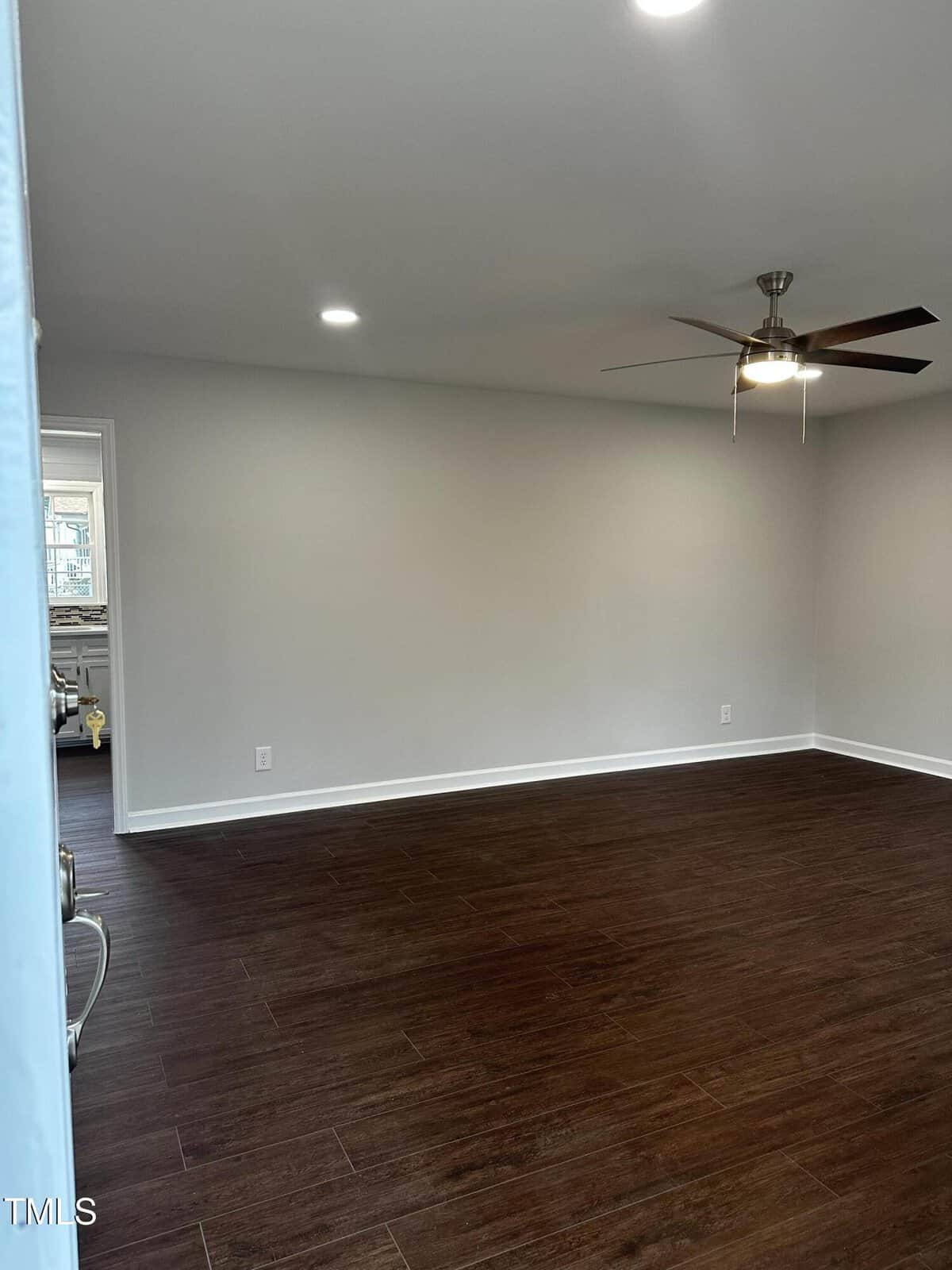 An empty room with dark wooden flooring and white walls. A ceiling fan with a light is centered on the ceiling. A window is partially visible through an open door on the left side of the room.