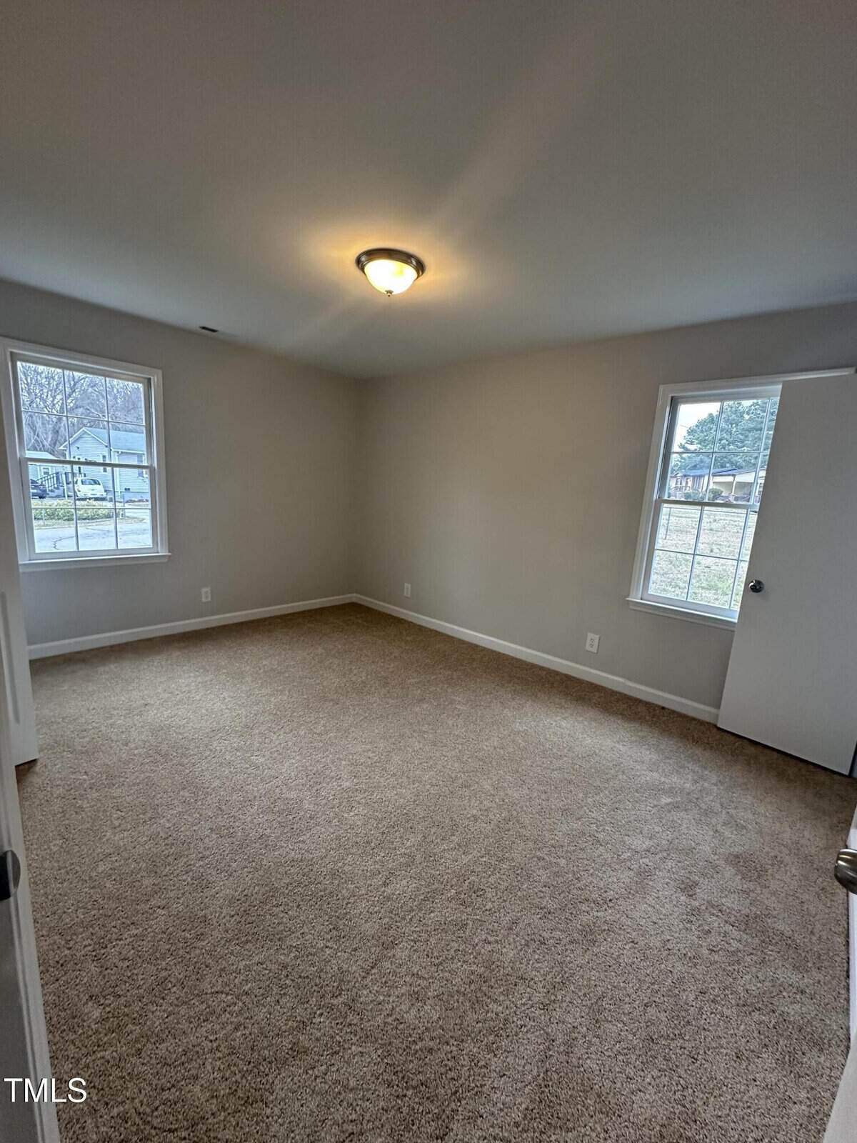 An empty room with beige carpet and gray walls. The ceiling features a light fixture. Two windows provide natural light, and there is a closed white door on the right.