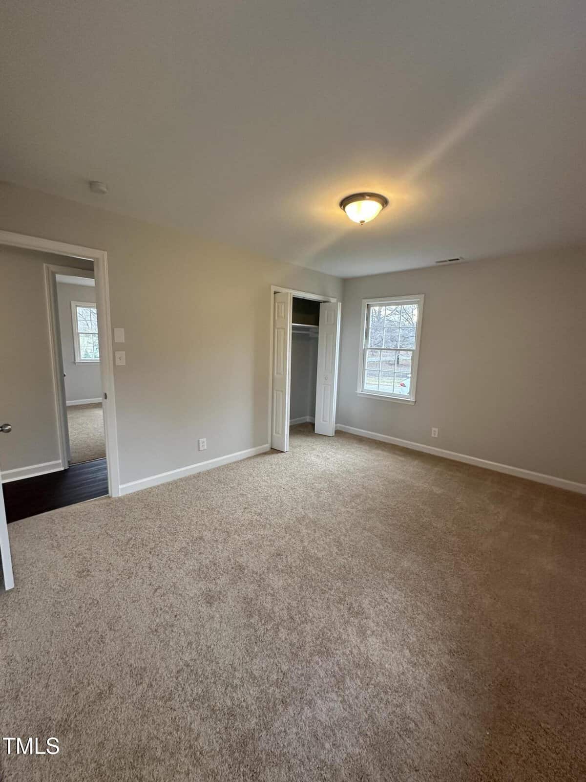 A carpeted room with beige walls, a ceiling light, and two windows. There is an open doorway leading to another room and a partially open door revealing a closet.