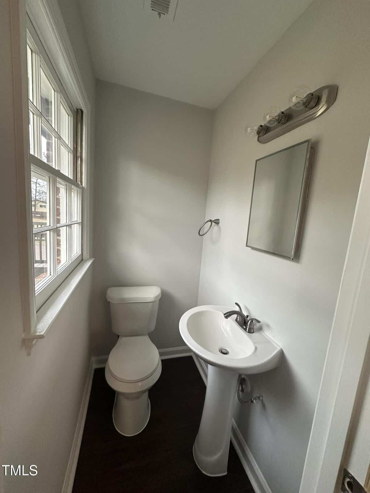 A small bathroom with a white pedestal sink and a toilet. A rectangular mirror is above the sink, and a light fixture with two bulbs is above the mirror. A window on the left wall allows natural light to enter the room.