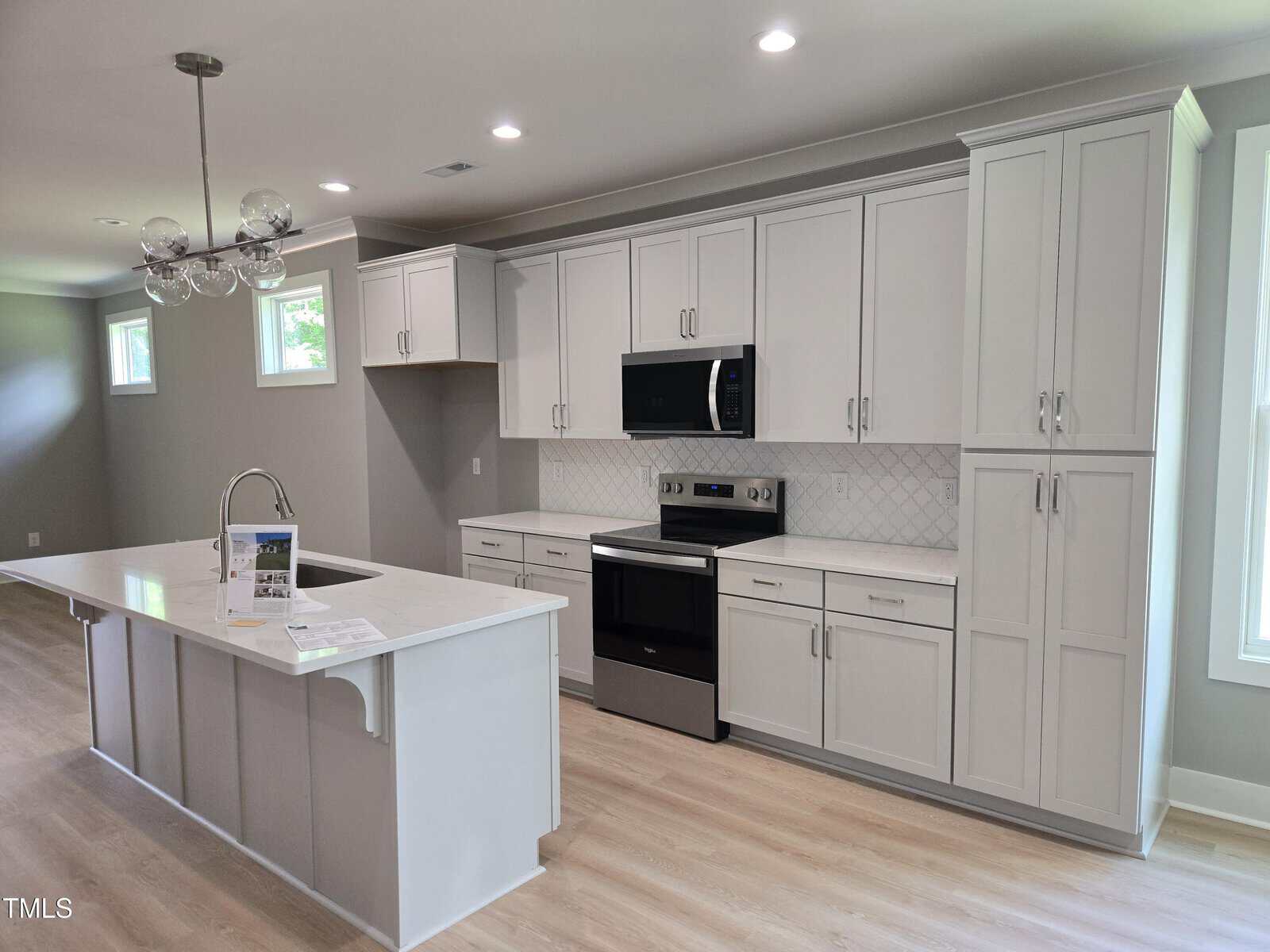 Modern kitchen with white cabinets, stainless steel appliances, an island with a sink, light wood flooring, and recessed lighting. A window allows natural light in. A brochure sits on the island countertop.