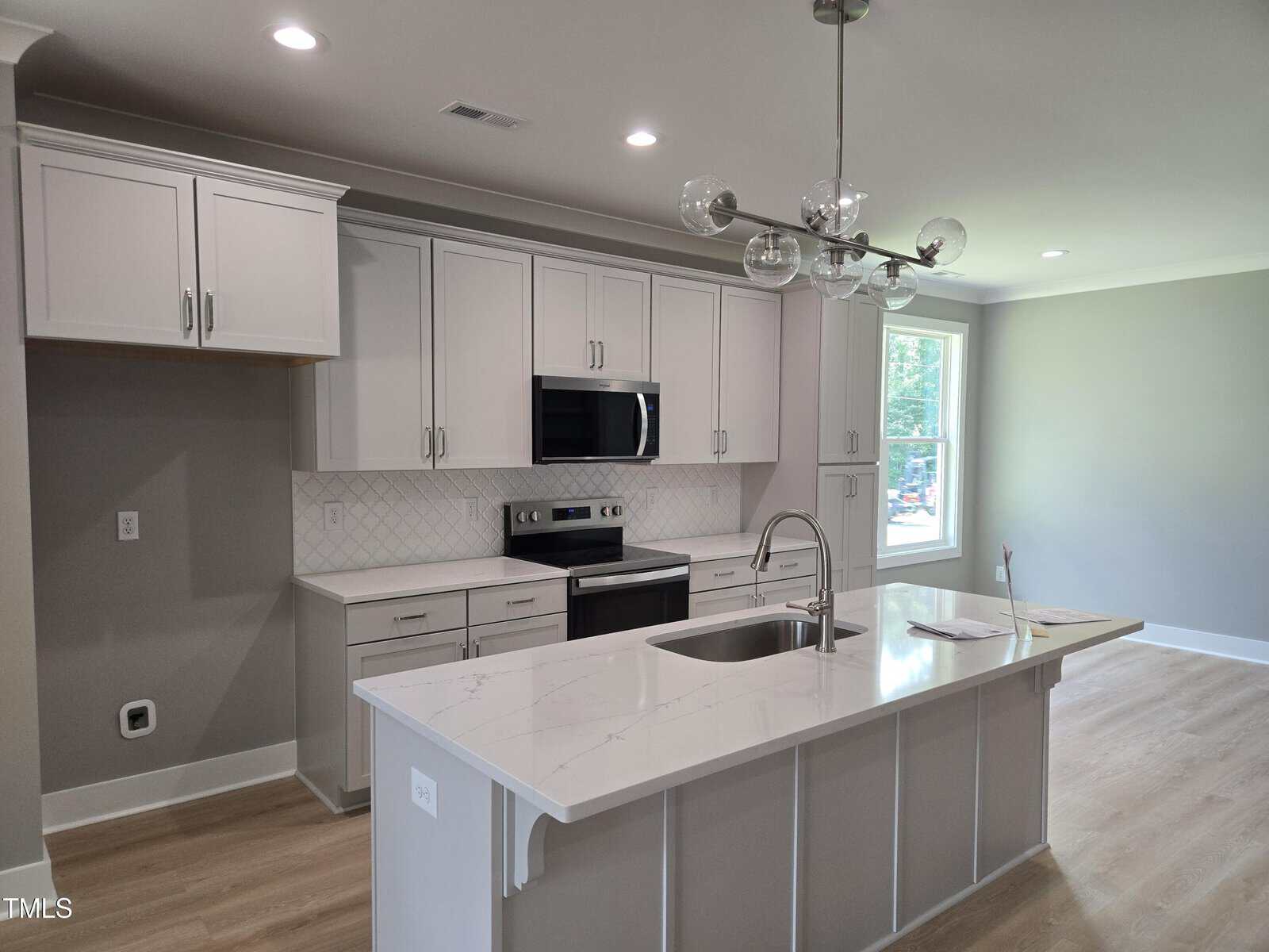 Modern kitchen with white cabinets, stainless steel appliances, an island with a sink and faucet, pendant lighting, light wood flooring, and a large window letting in natural light.