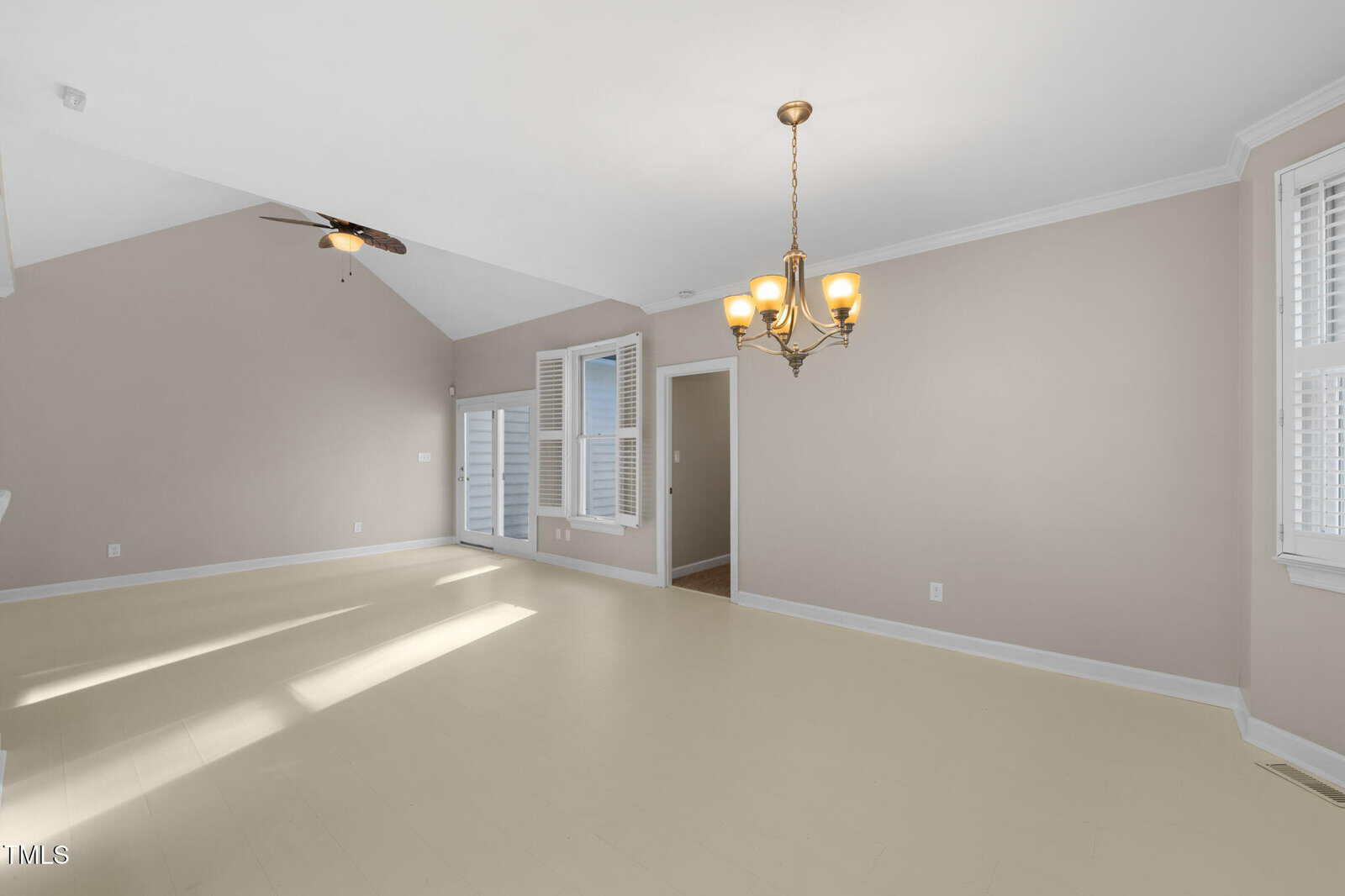 A spacious empty room with beige walls and light-colored flooring. It features a ceiling fan, a chandelier, and several windows with white shutters. A door leads to another room, and sunlight is streaming in through the windows.