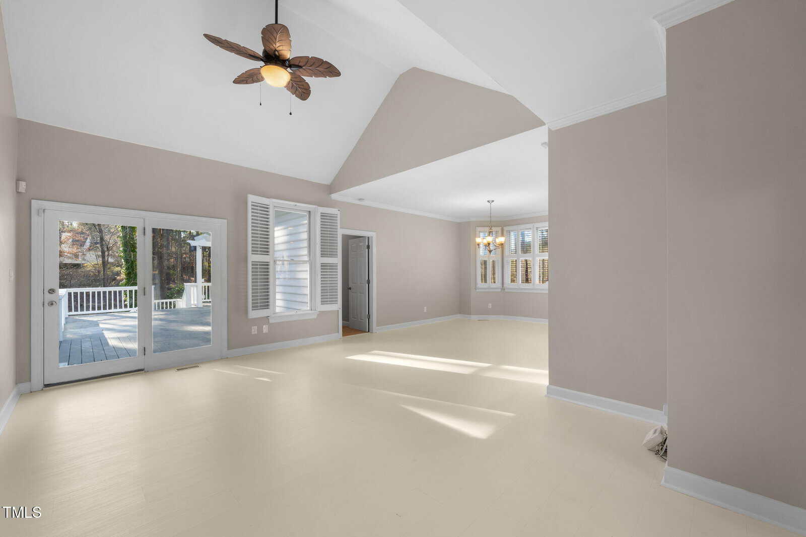 A spacious living room with light-colored walls and a vaulted ceiling features a ceiling fan and a large window. Glass doors open to a deck outside. The room has hardwood floors and a small dining area with a chandelier near another set of windows.