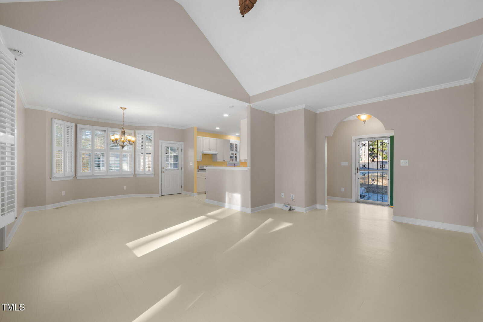 A spacious empty room features light beige walls and floors, large windows, and an open doorway leading to a kitchen area. A chandelier hangs from the ceiling, and a glass door opens to a patio outside.