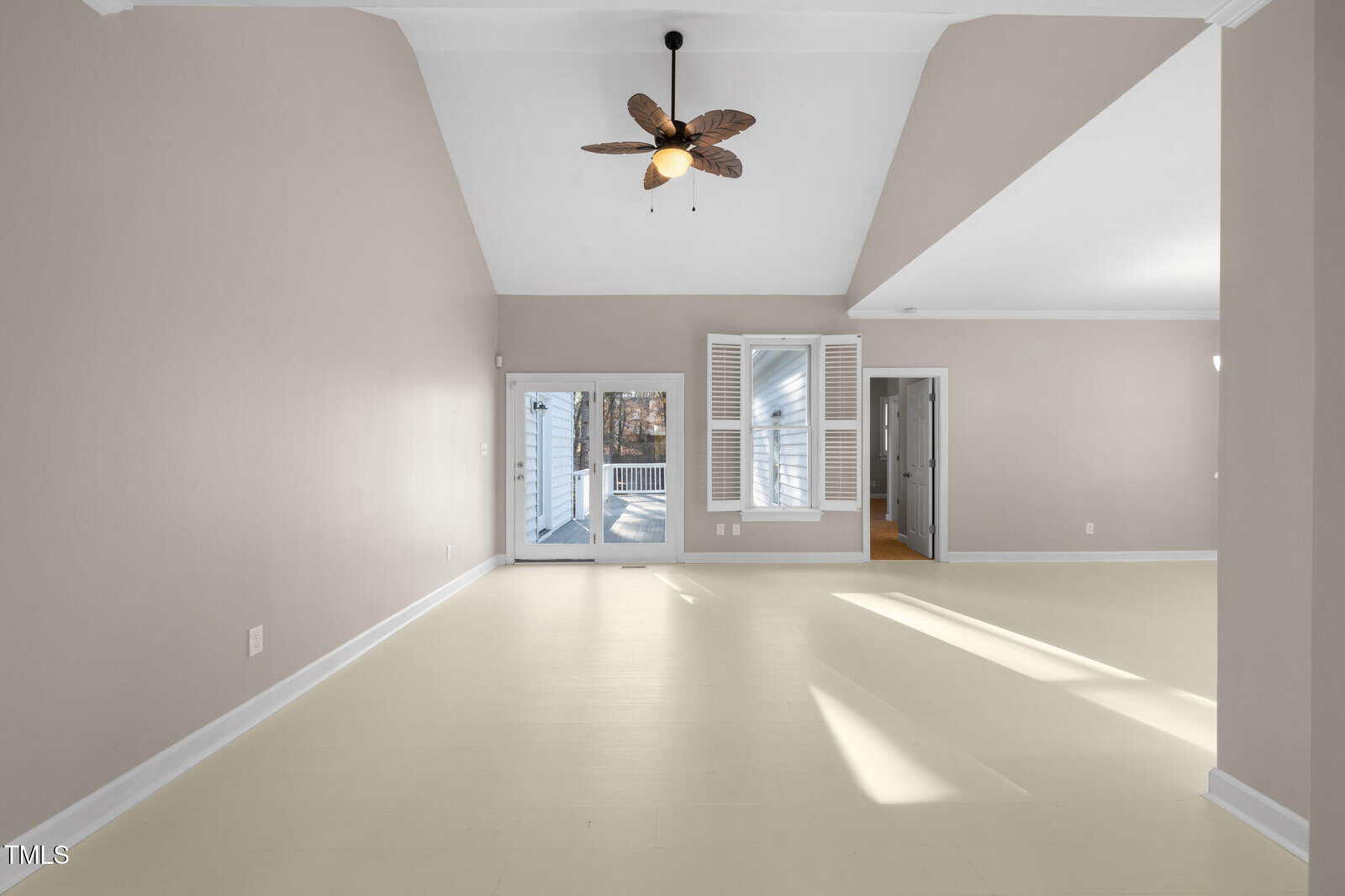 A spacious, empty room with light beige walls and a high ceiling featuring a decorative fan. Sunlight streams through large glass doors that lead to an outdoor deck. There is a window with shutters and an open doorway to another room.