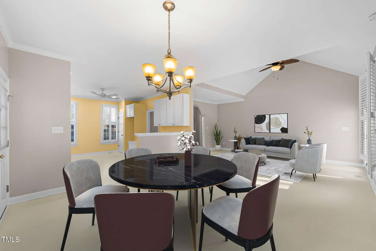 A dining area with a round, black table and six chairs is in the foreground. A chandelier hangs above. In the background, there's a living area with a gray sofa, artwork on the wall, and a ceiling fan. The open layout features light walls and flooring.