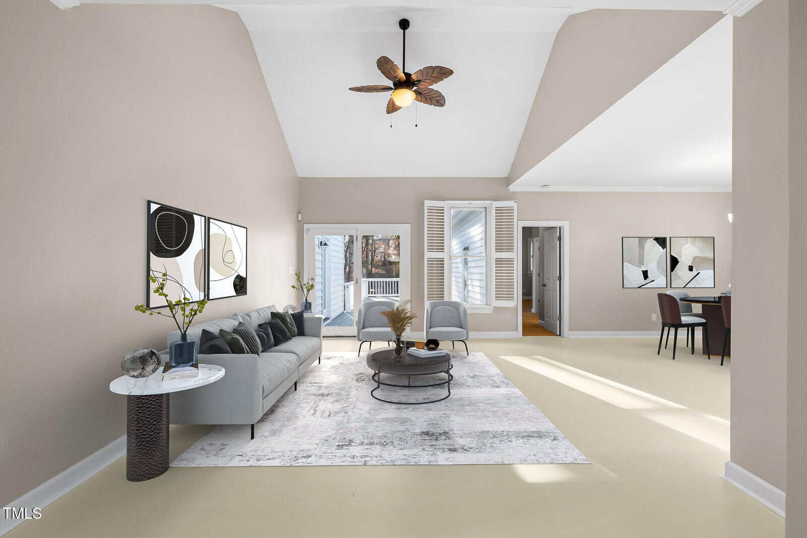 A spacious living room with beige walls, featuring a gray sofa, modern art on the wall, and a ceiling fan. A coffee table and plants decorate the room. Large windows let in natural light, and there is a view of an adjacent room with a dining table.