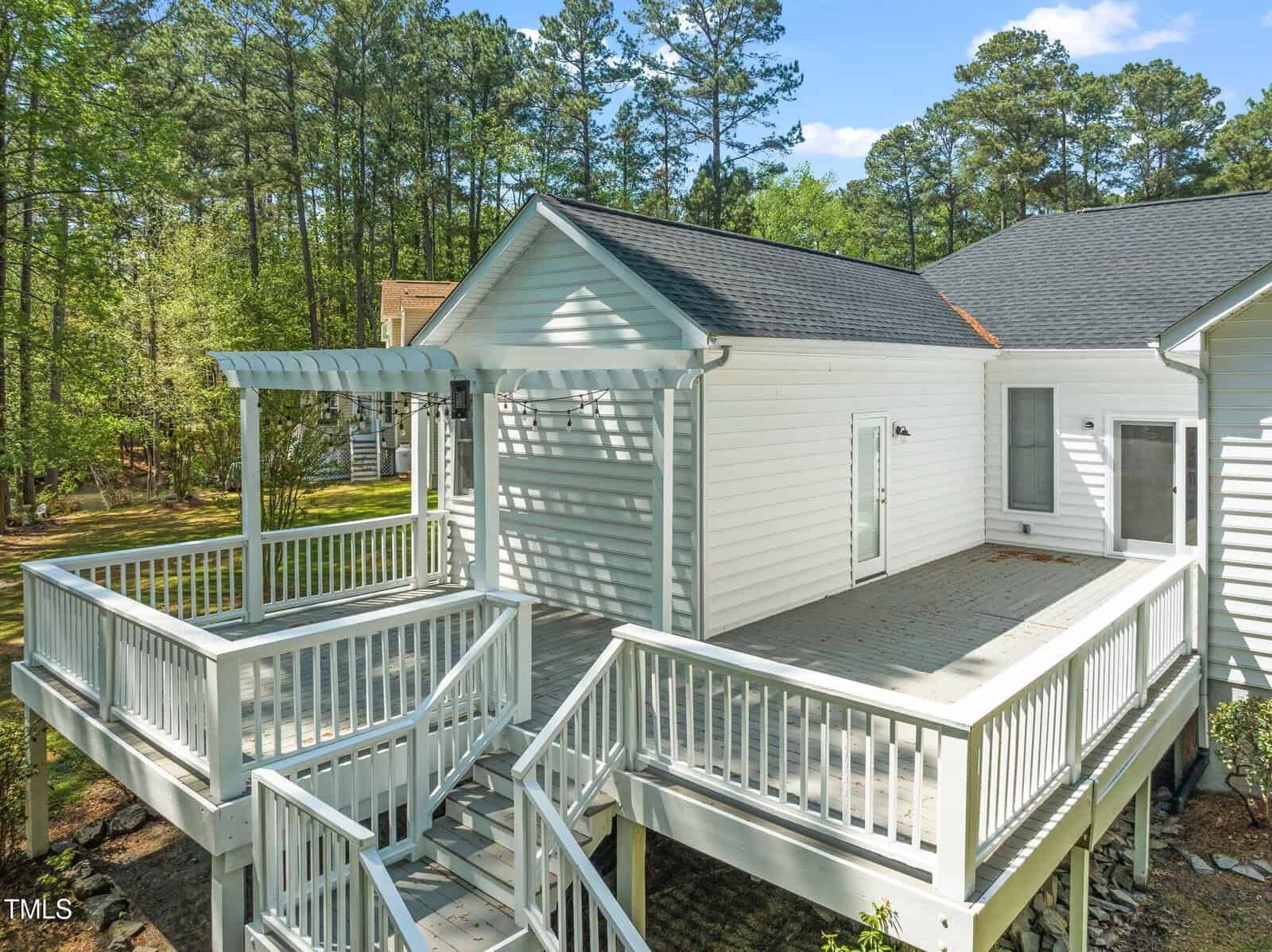 A white wooden house with a large elevated deck featuring railings, stairs, and a pergola. The deck is surrounded by trees and greenery, and the house has a dark shingle roof and white siding.