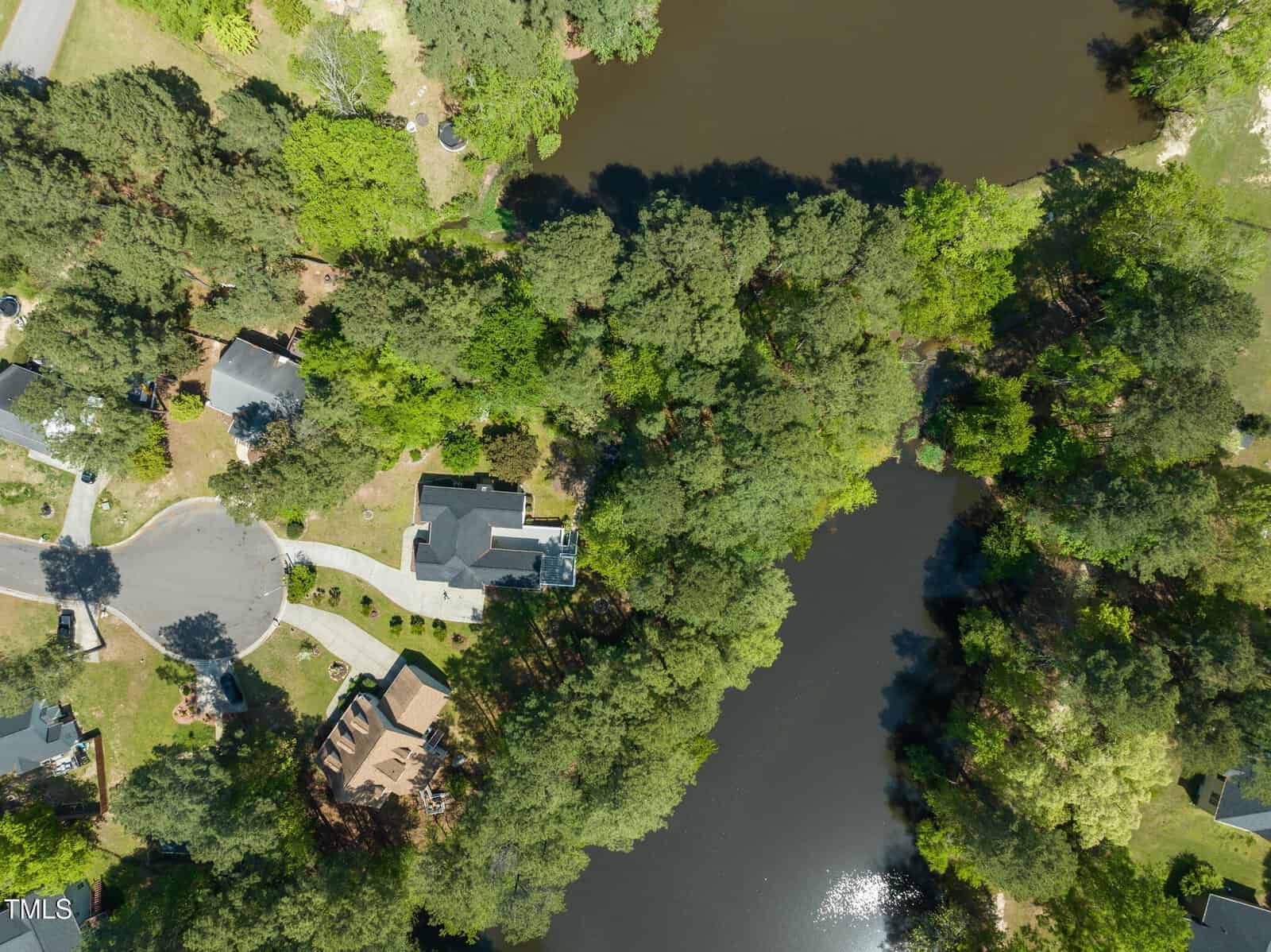 Aerial view of a suburban neighborhood featuring several houses surrounded by dense trees and greenery, with two small bodies of water partially visible in the upper and lower portions of the image.