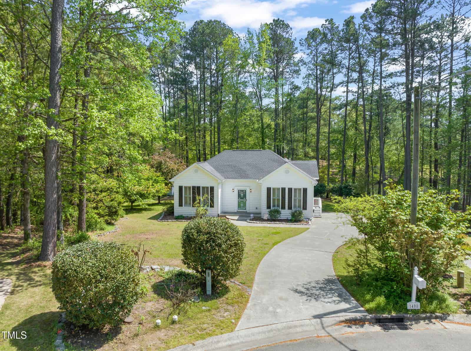A single-story white house with black shutters at 1408 Ferrell Court in Creedmoor is surrounded by trees. A curved driveway leads up to the front door, with bushes and grass in the yard, all set against a wooded backdrop.