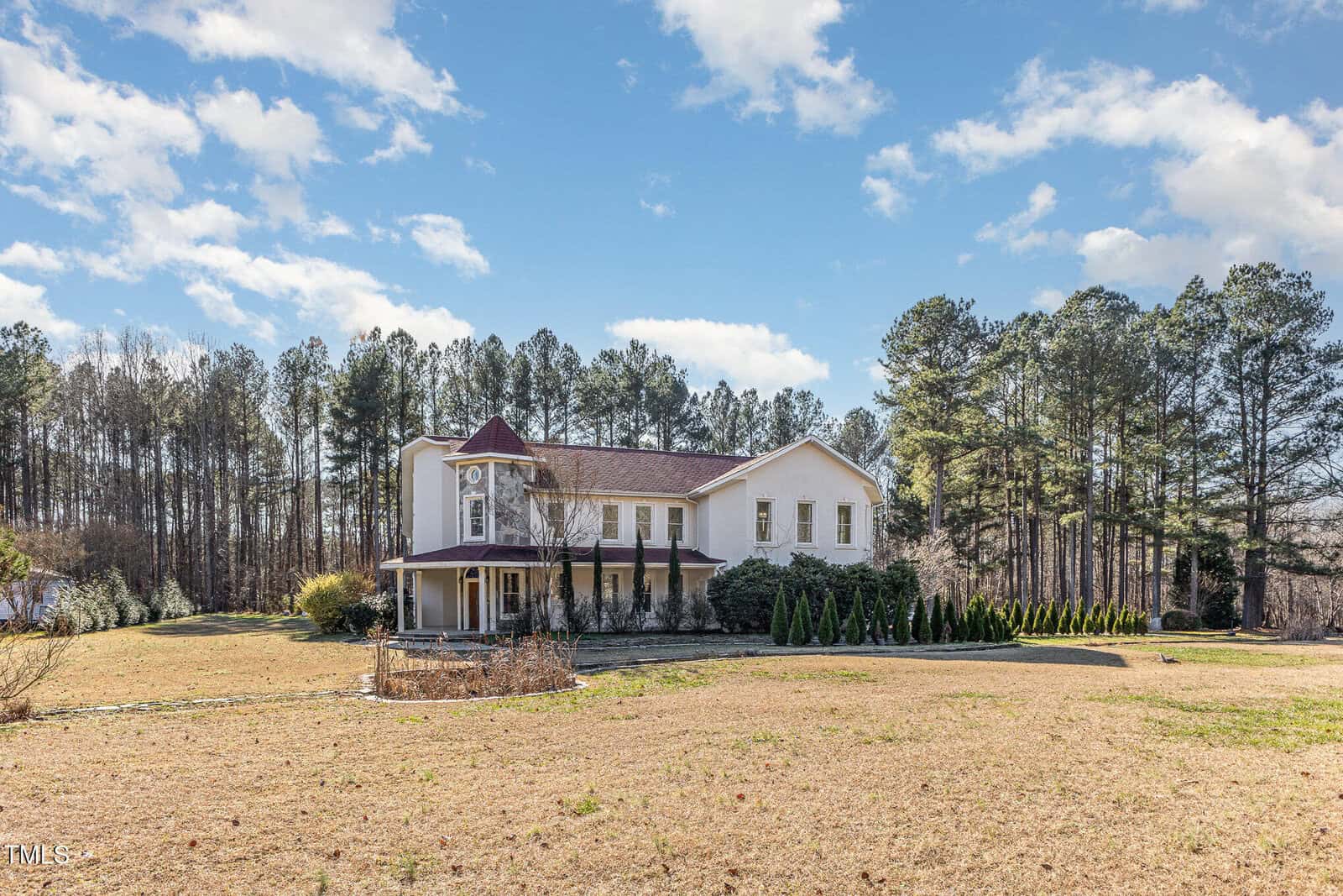 A large two-story white house with a wraparound porch and red roof sits on a spacious, grassy lot with tall trees in the background under a blue sky with scattered clouds.