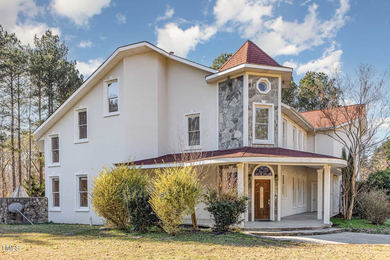 Two-story house with stucco walls and a turret featuring stone accents, a sloped red roof, covered entryway with columns, arched wooden door, and landscaped bushes in the front yard on a clear day.