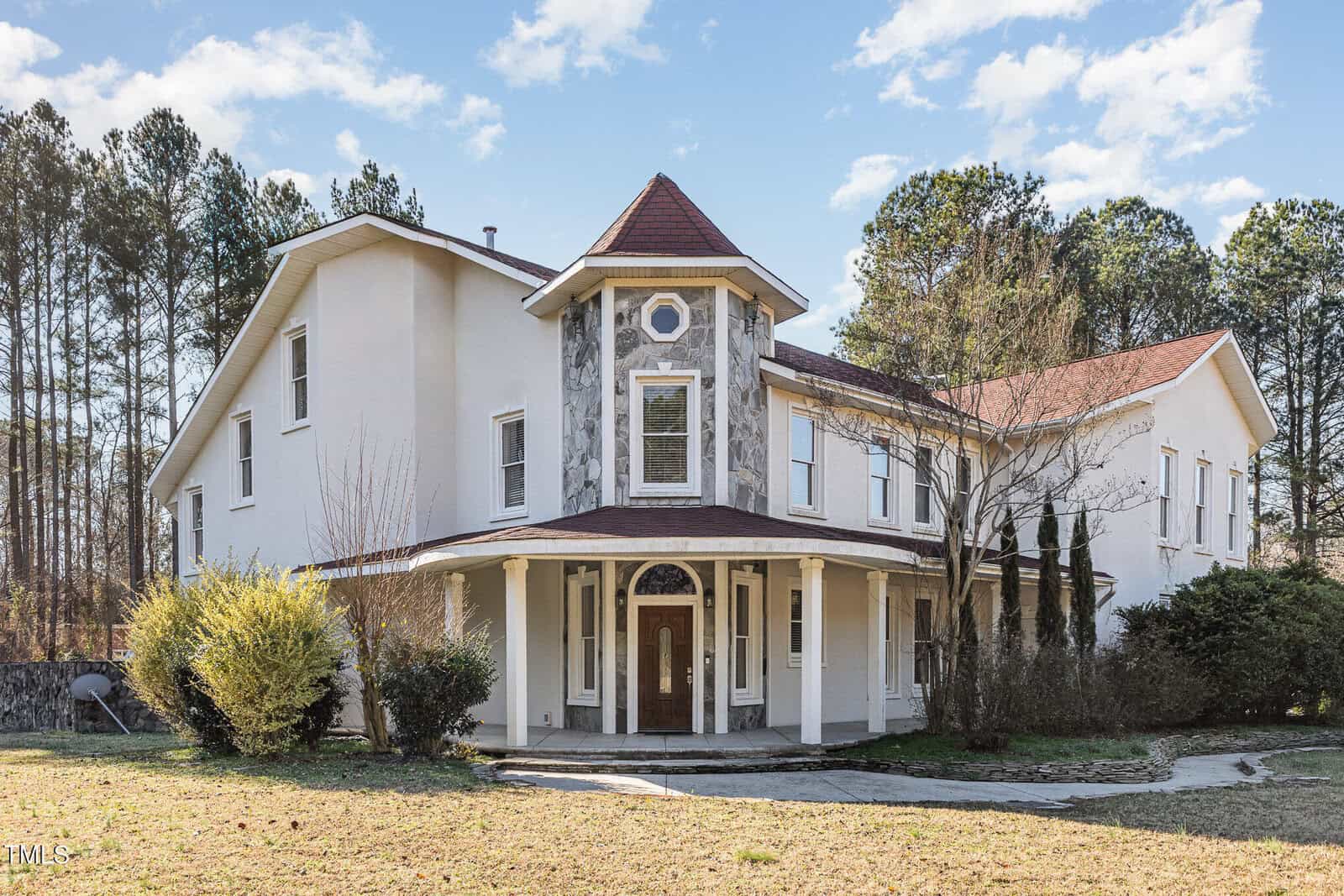 A large, two-story house with white exterior walls, a prominent turret, red roof, and wraparound porch. The house is surrounded by trees and bushes, with a mostly bare lawn in front.