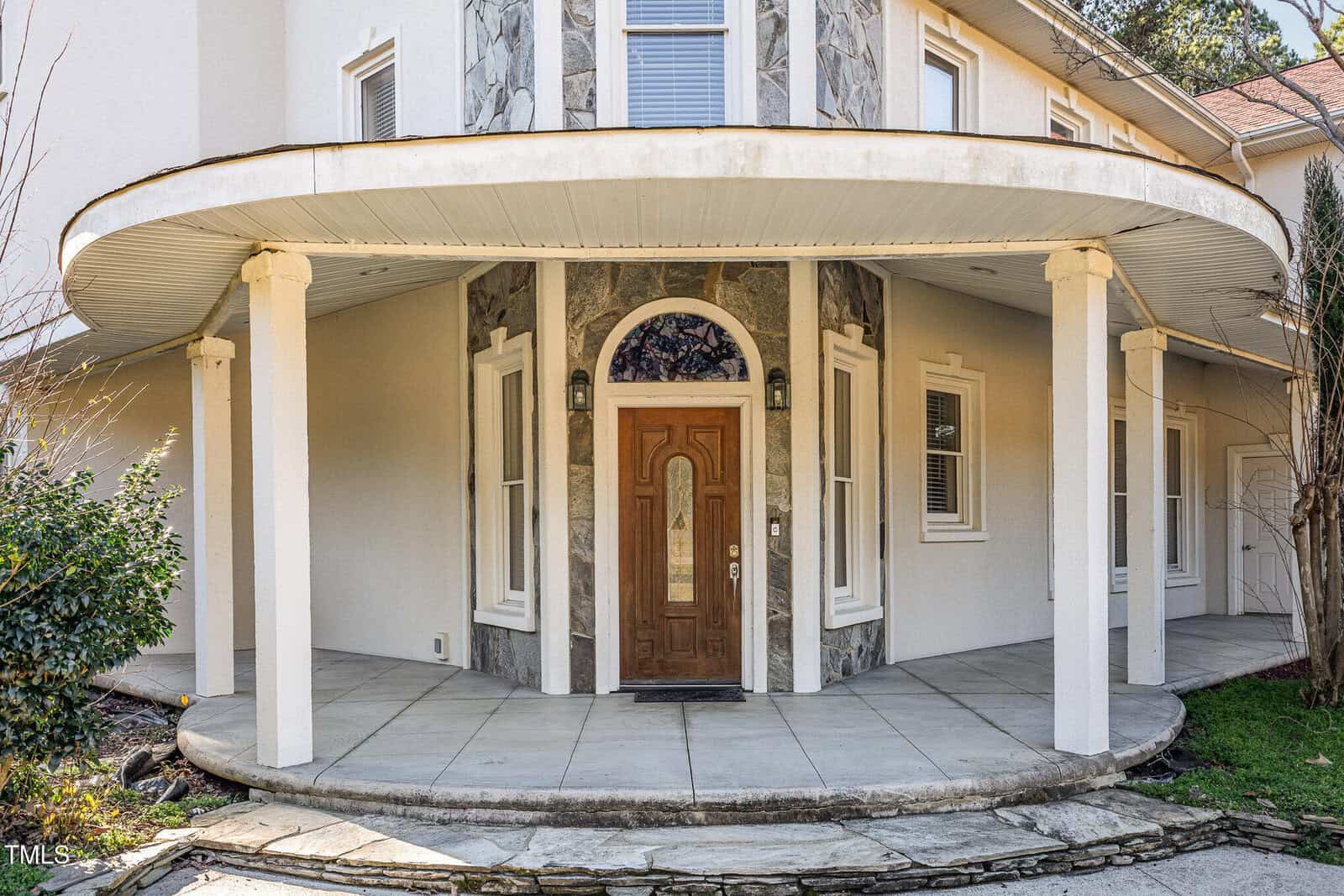 A house entrance featuring a wooden door with a glass insert, surrounded by stone accents and white stucco walls. The entryway is covered by a curved porch roof supported by white columns.