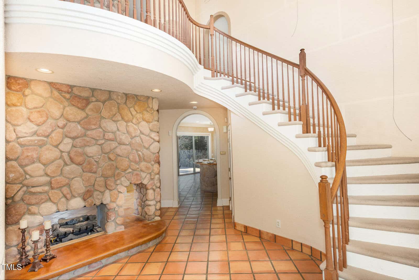 A curved staircase with wooden railings is next to a stone fireplace. The floor is covered with terracotta tiles, and an arched doorway leads to another room with similar flooring.